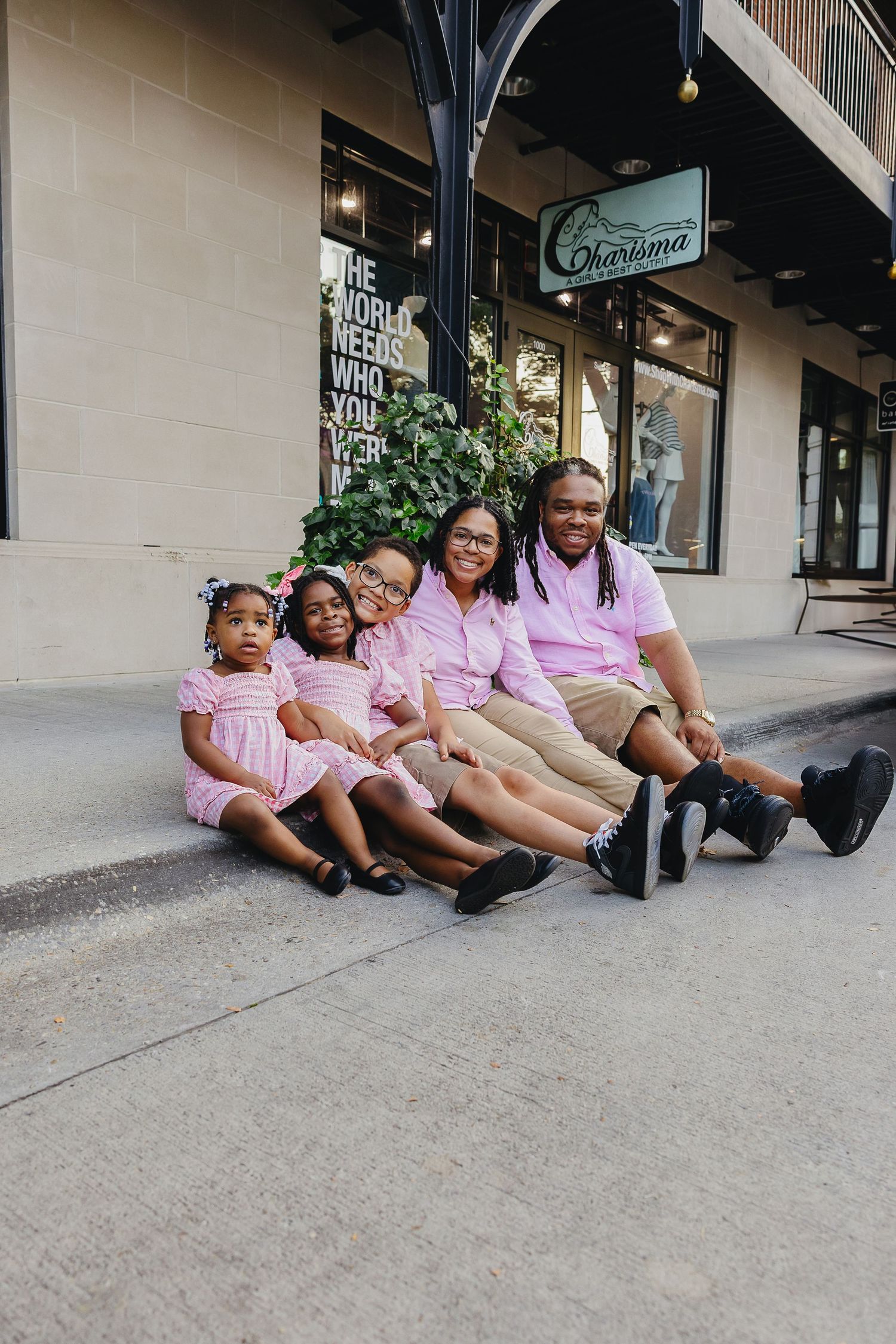 Happy family of five in coordinating pink outfits sitting together on sidewalk outside boutique.