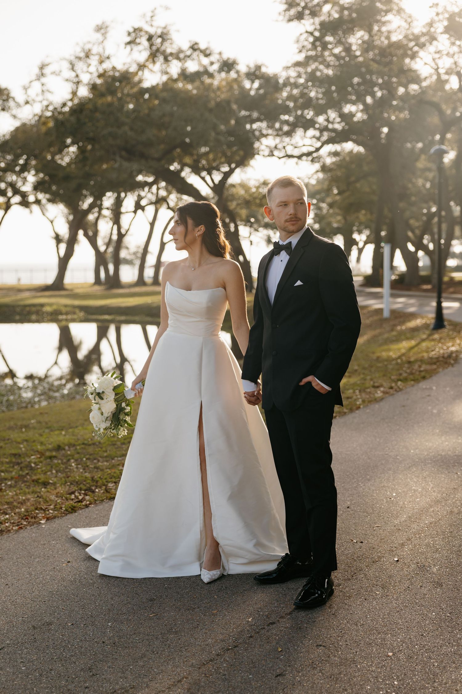 Elegant bride in strapless white gown and groom in black tuxedo pose on a sunlit outdoor path.