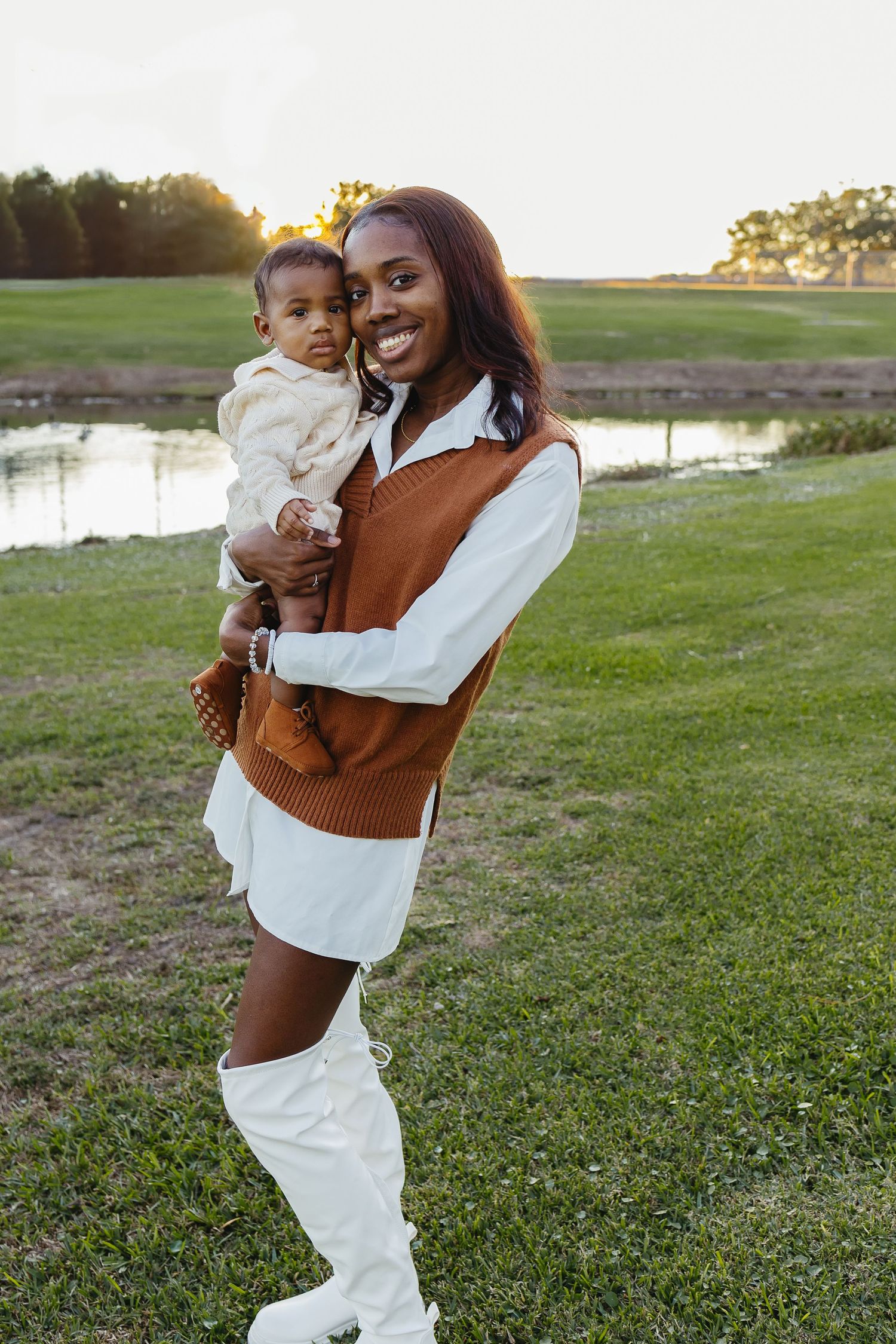 Smiling Black mother holding baby outdoors at sunset, wearing brown vest over white dress with white knee boots.