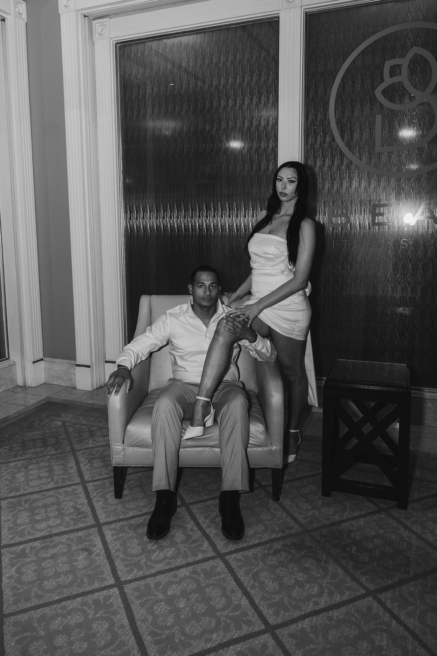Black and white photo of couple posing indoors, man seated in armchair while woman in white dress stands beside him.