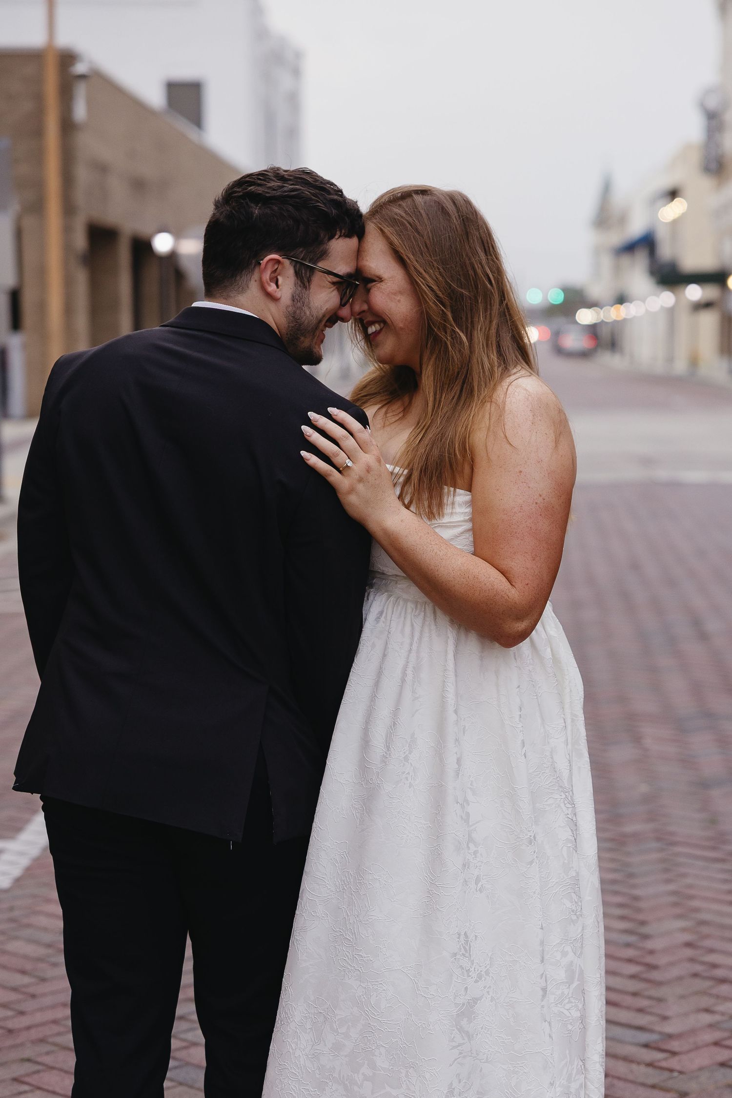 Newlywed couple touching foreheads and laughing on brick street at dusk, bride in white lace dress and groom in black suit.