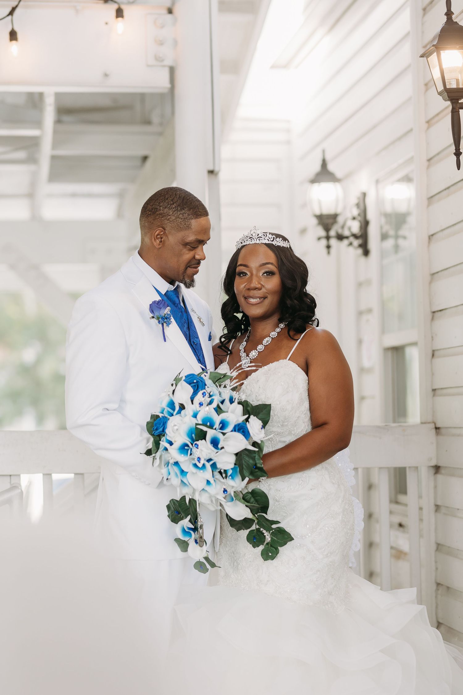 Smiling bride in tiara holds blue calla lily bouquet with groom in white suit outdoors.