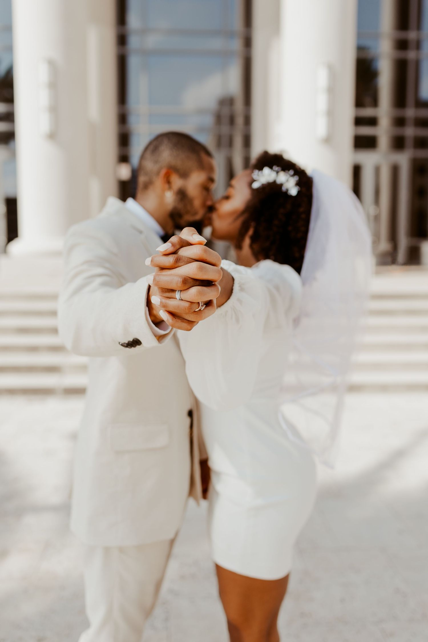 Bride and groom in white attire sharing first dance kiss outside elegant columned building.