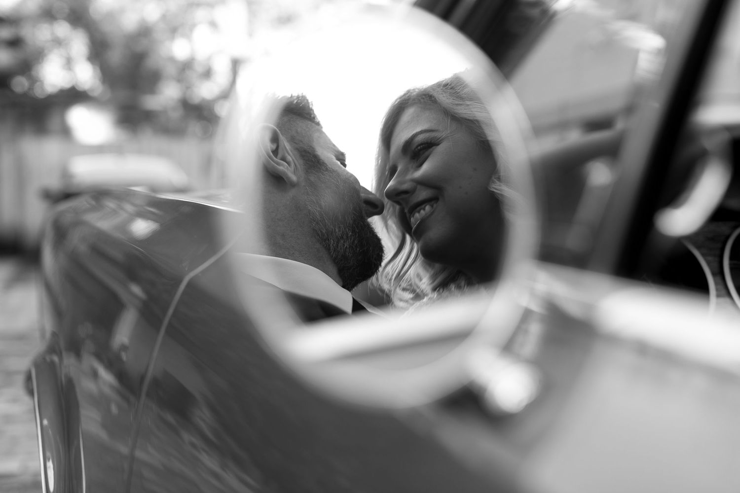 Black and white reflection of smiling bride and groom sharing intimate moment in car mirror.