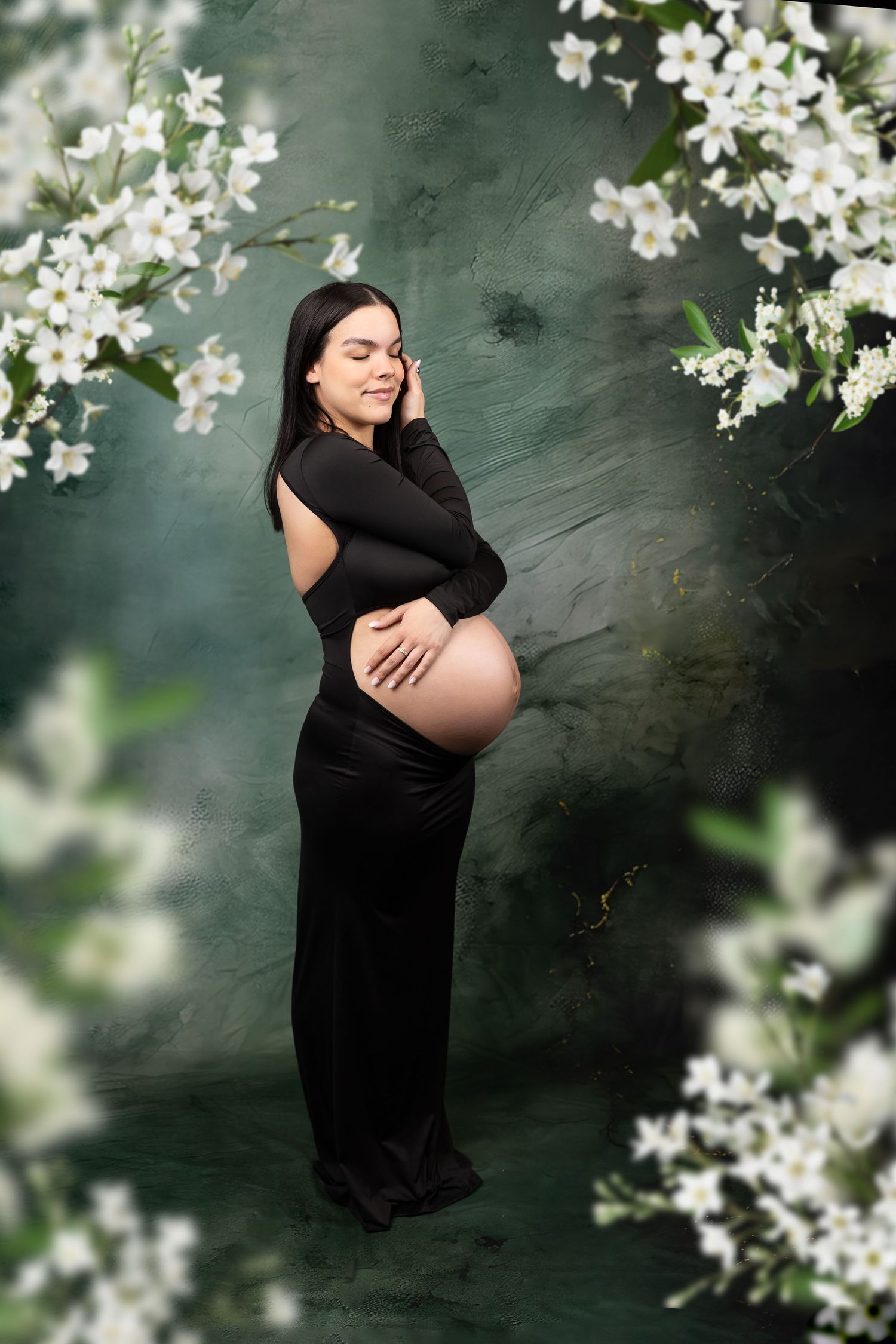 Smiling pregnant woman in black dress surrounded by white flowers on a green backdrop.