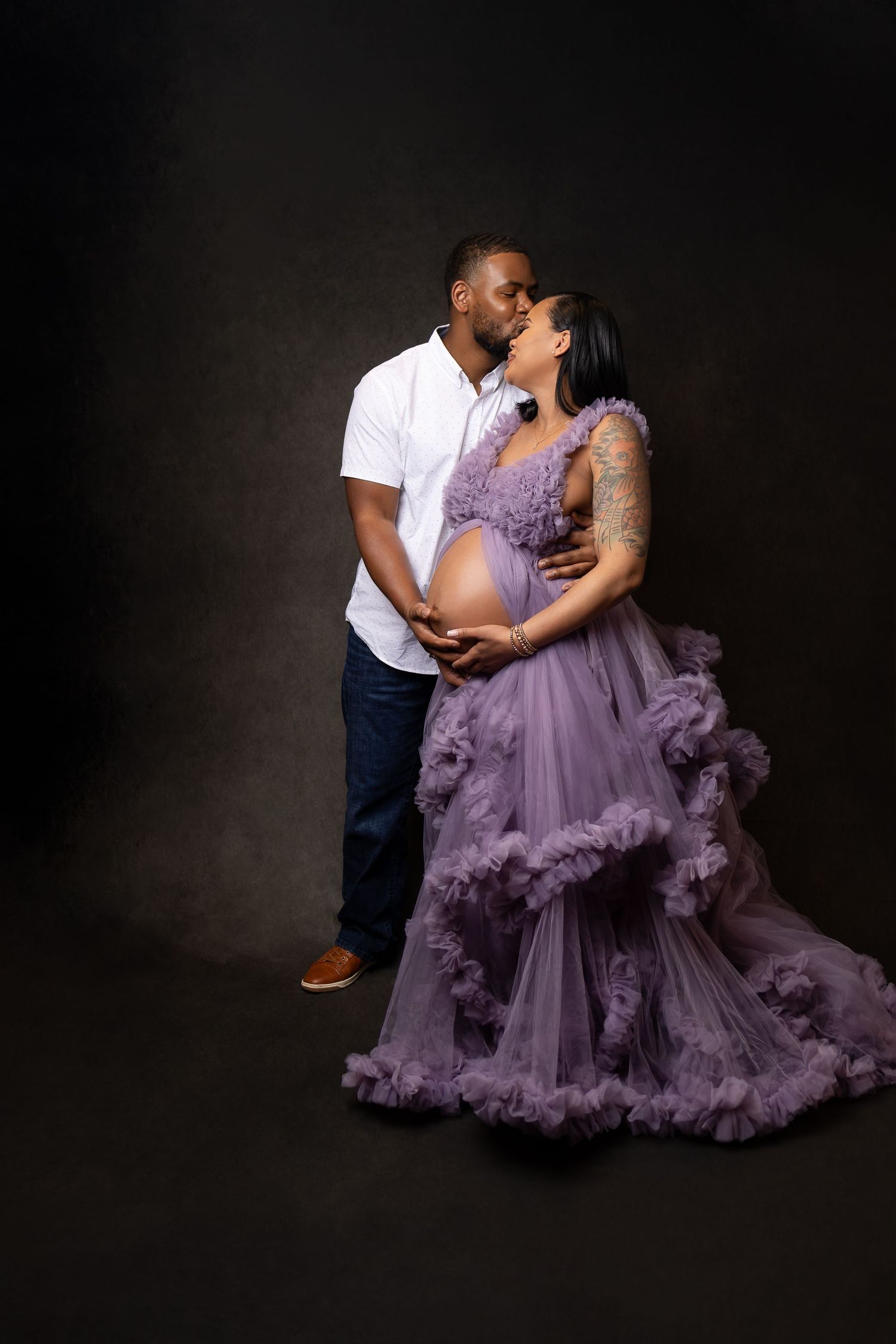 Couple sharing a kiss during maternity photoshoot, woman in purple tulle gown cradling baby bump.