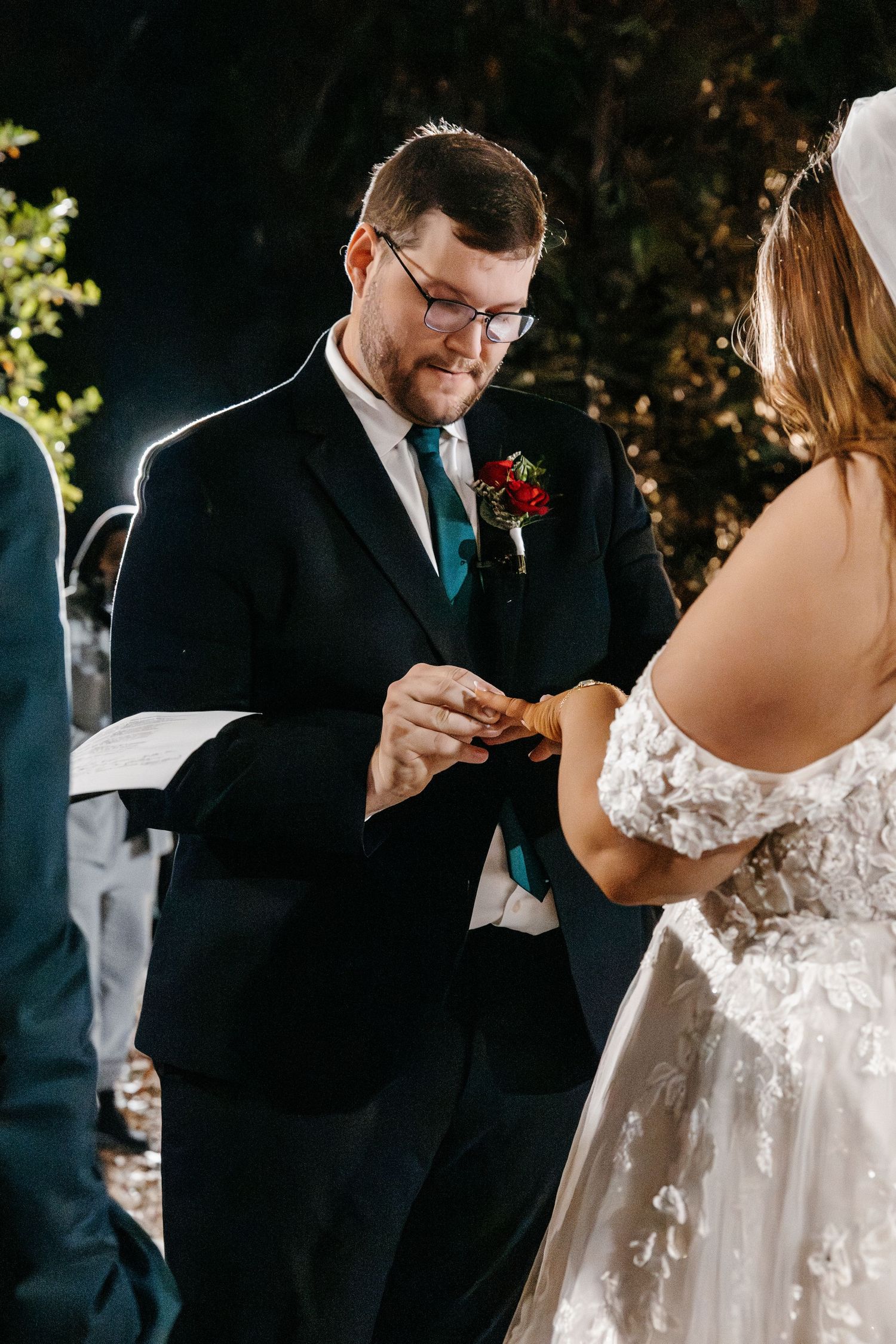 Groom placing wedding ring on bride's finger during outdoor night ceremony.