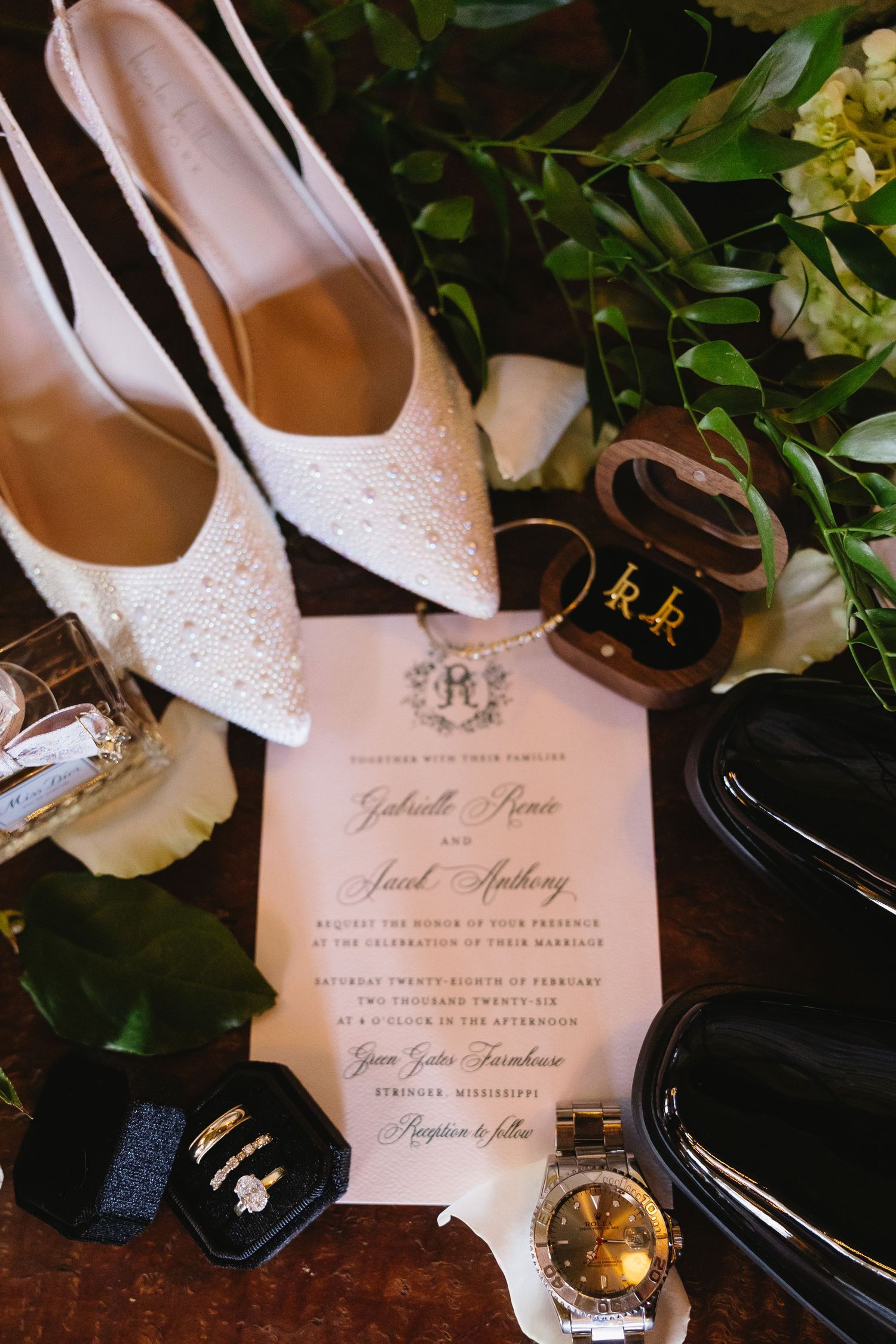 Wedding flat lay with lace heels, invitation, rings, watch, and white floral bouquet on a dark surface.