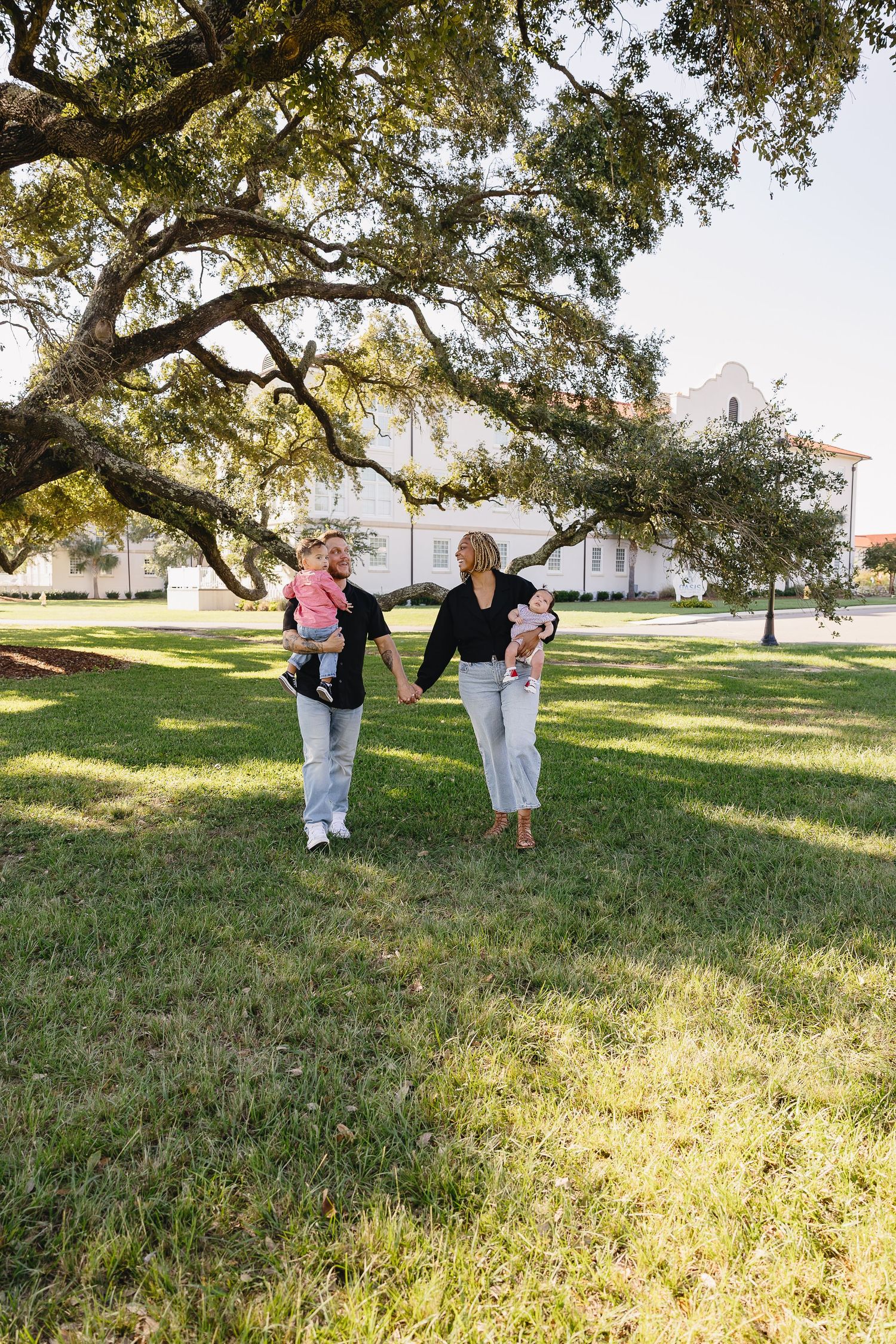 Two women holding young children walking hand in hand across green lawn beneath sprawling oak tree.