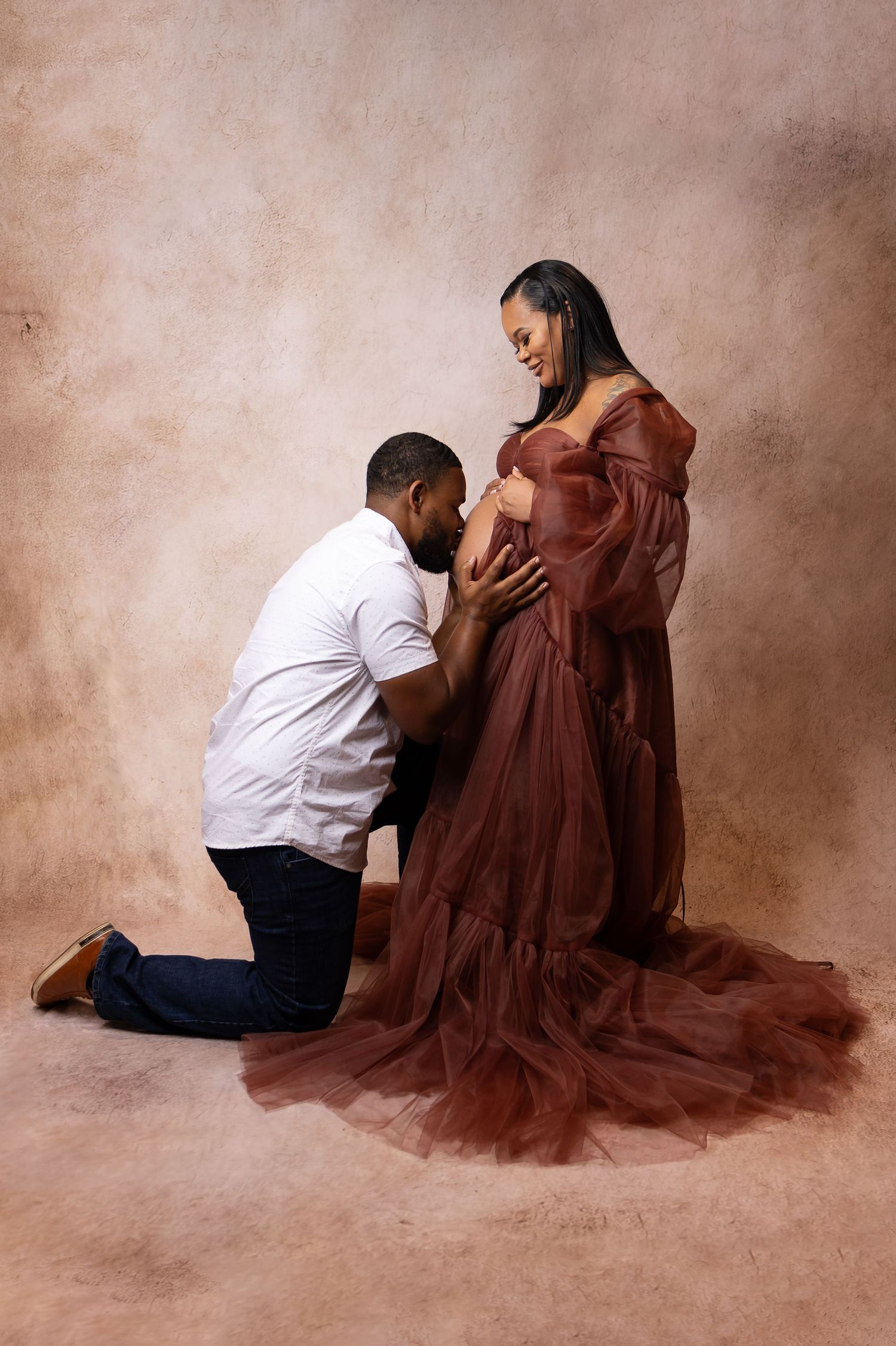 Man kneeling to kiss pregnant womans belly while she smiles in rust tulle gown.