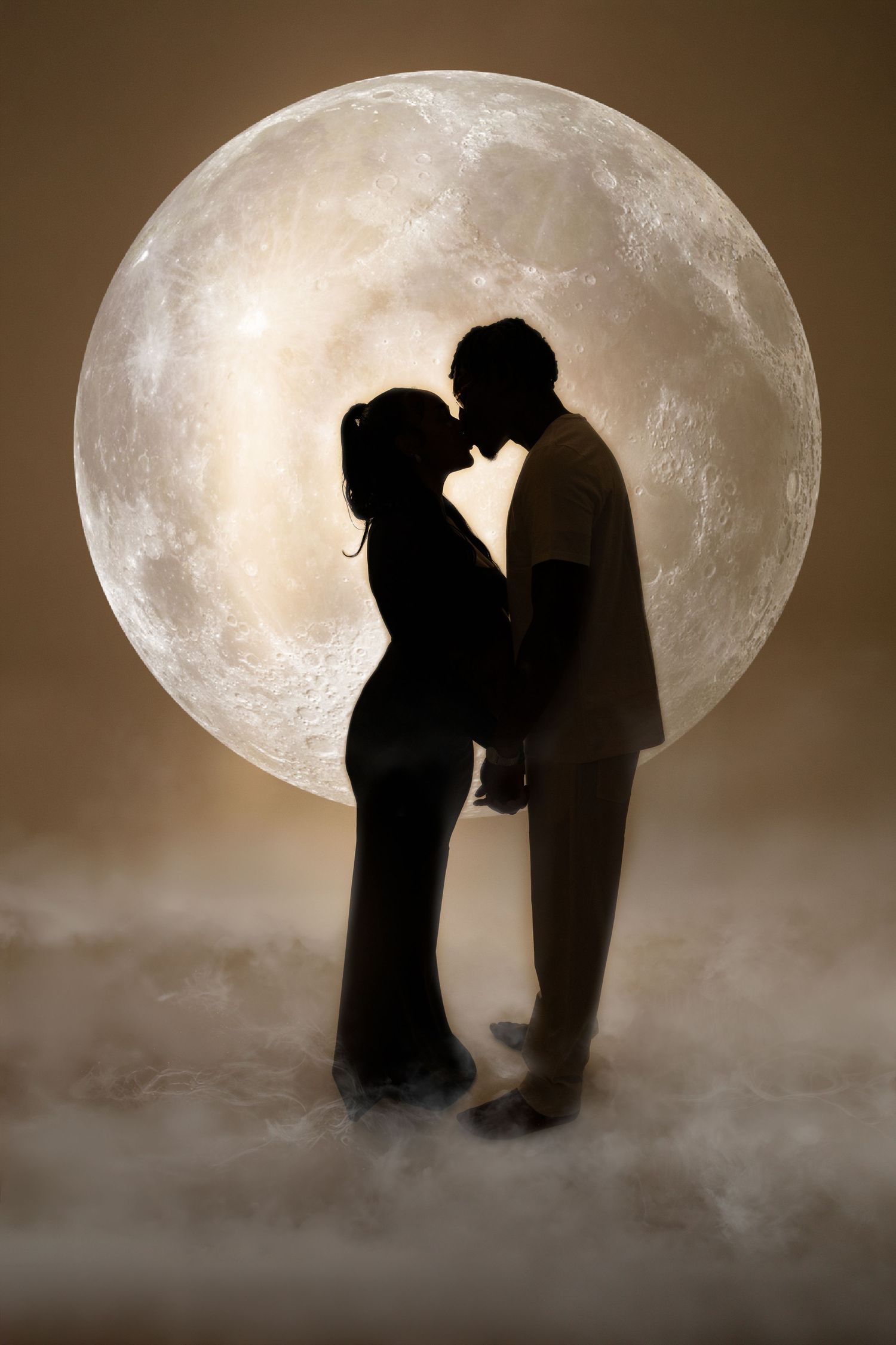 Silhouette of pregnant couple kissing in front of large full moon with misty foreground.