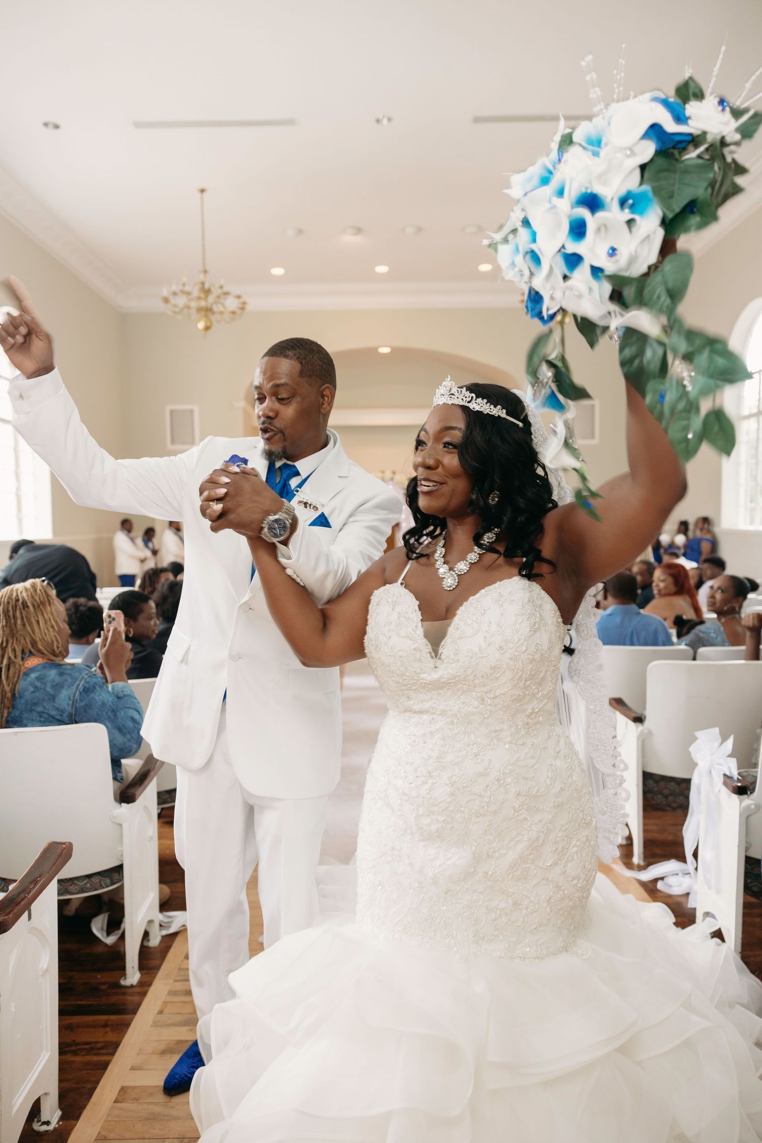 Excited bride raising blue and white bouquet as groom cheers while exiting wedding ceremony.