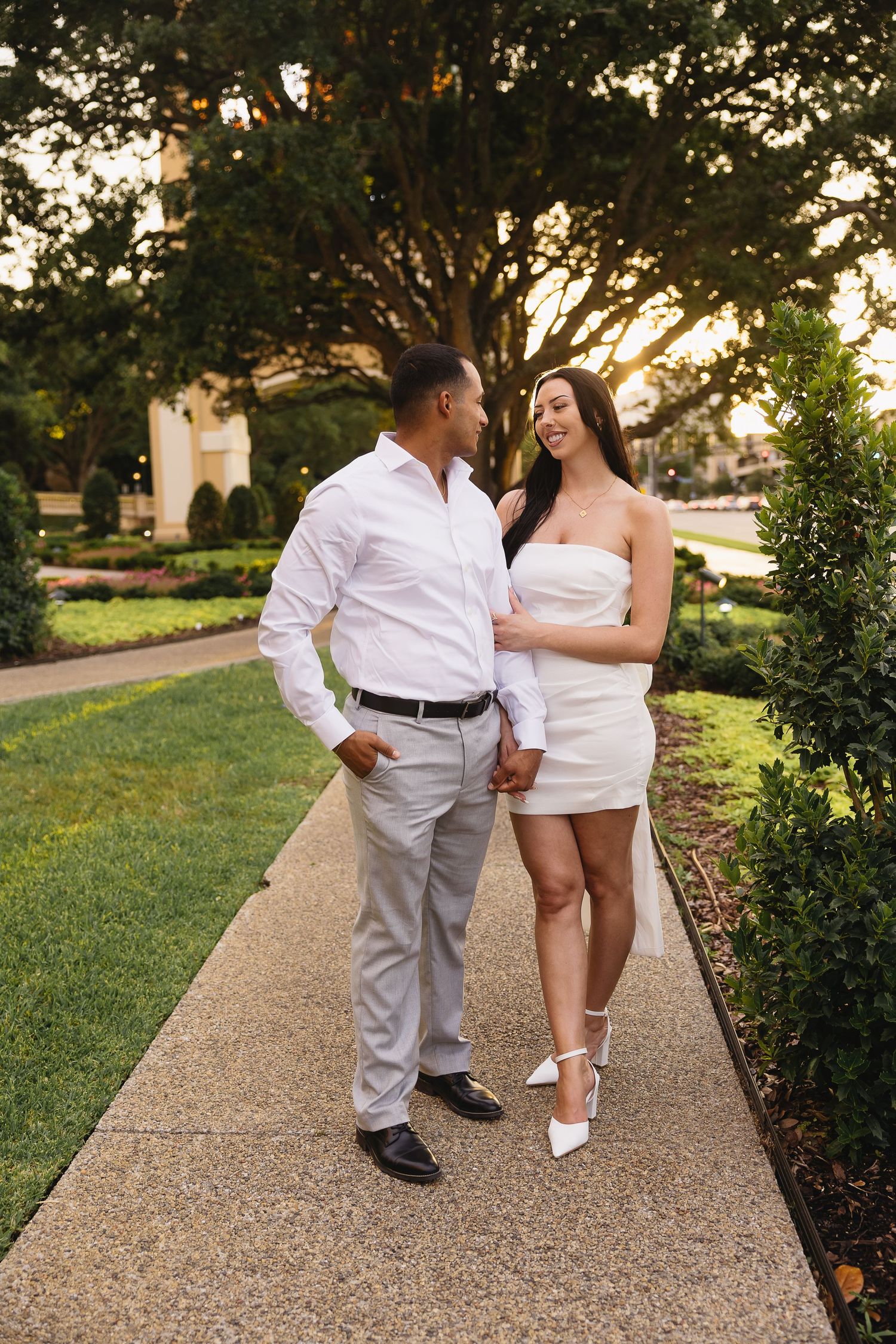 Happy couple walking together on garden path at golden hour, woman in white dress and man in white shirt smiling.