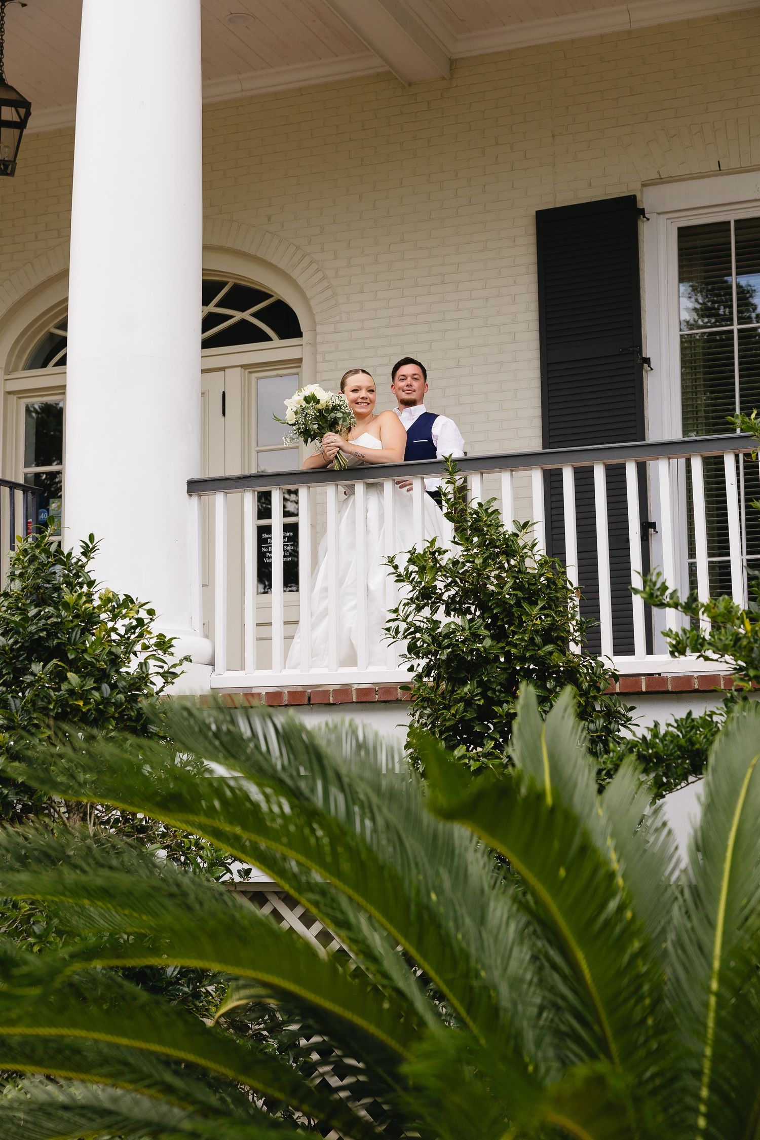 Bride and groom posing on a white colonial mansion balcony surrounded by lush greenery on their wedding day.