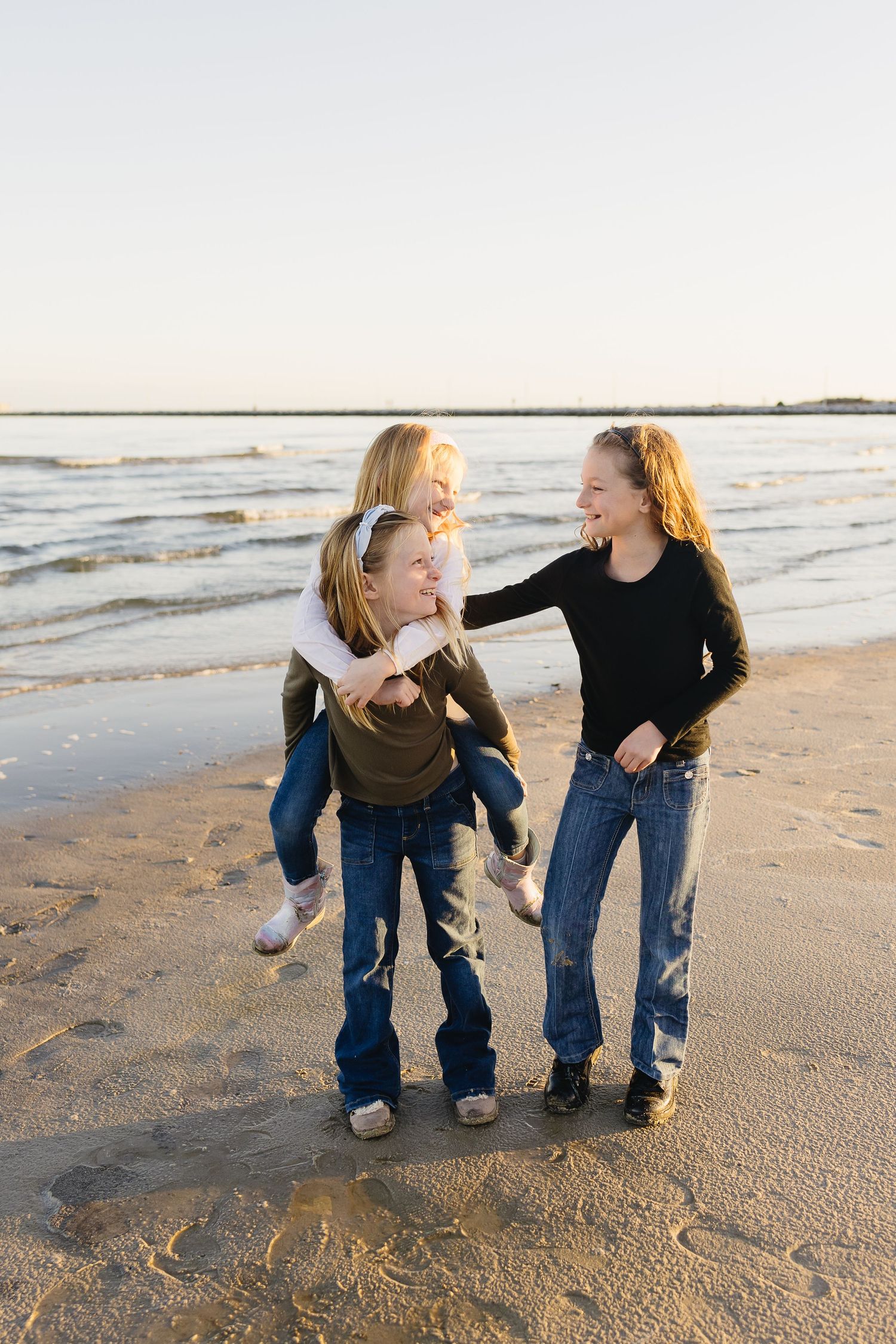 Three girls laugh and play piggyback together on a sandy beach at sunset.