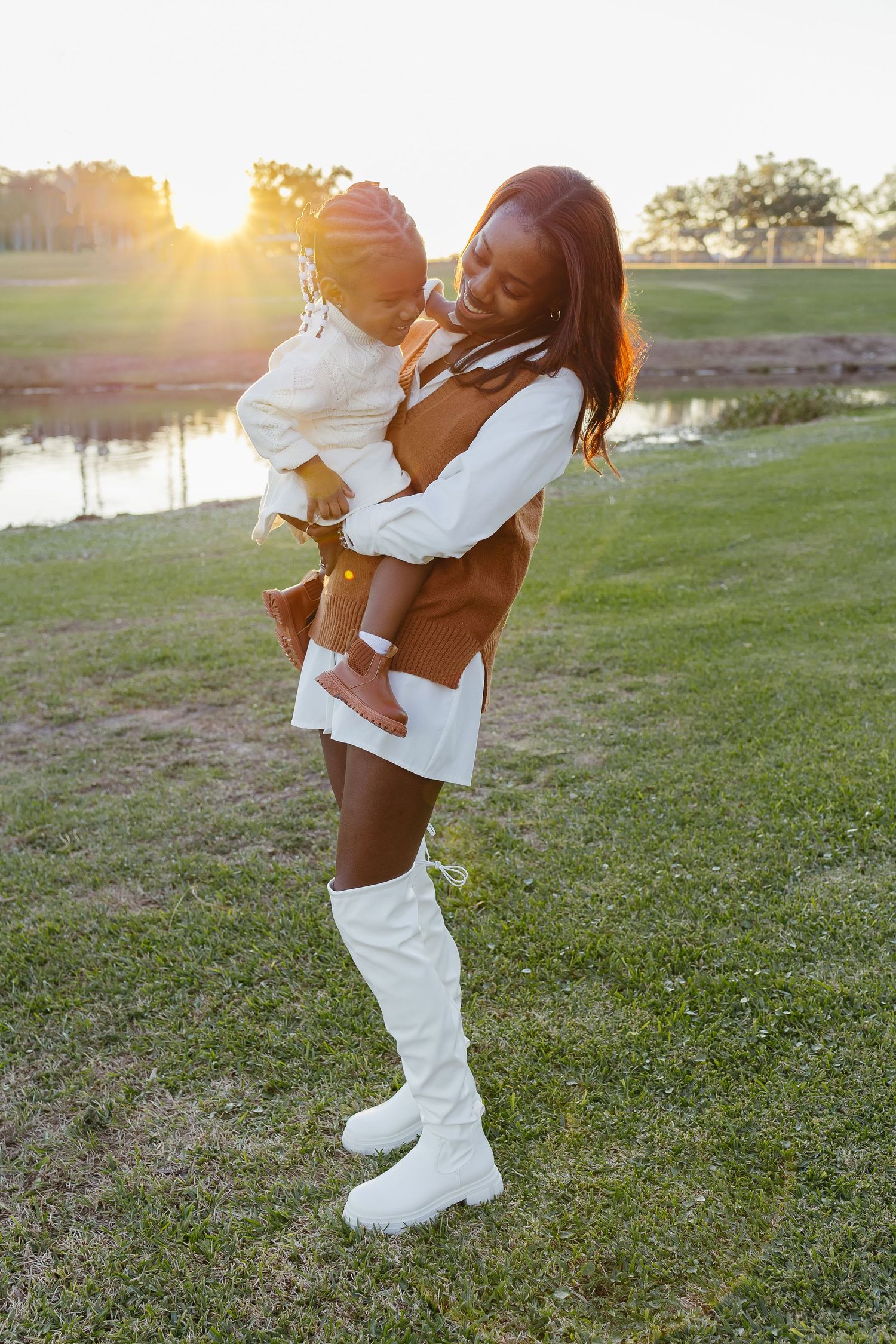 A mother holding her young daughter while smiling in a sunlit grassy park near a pond.