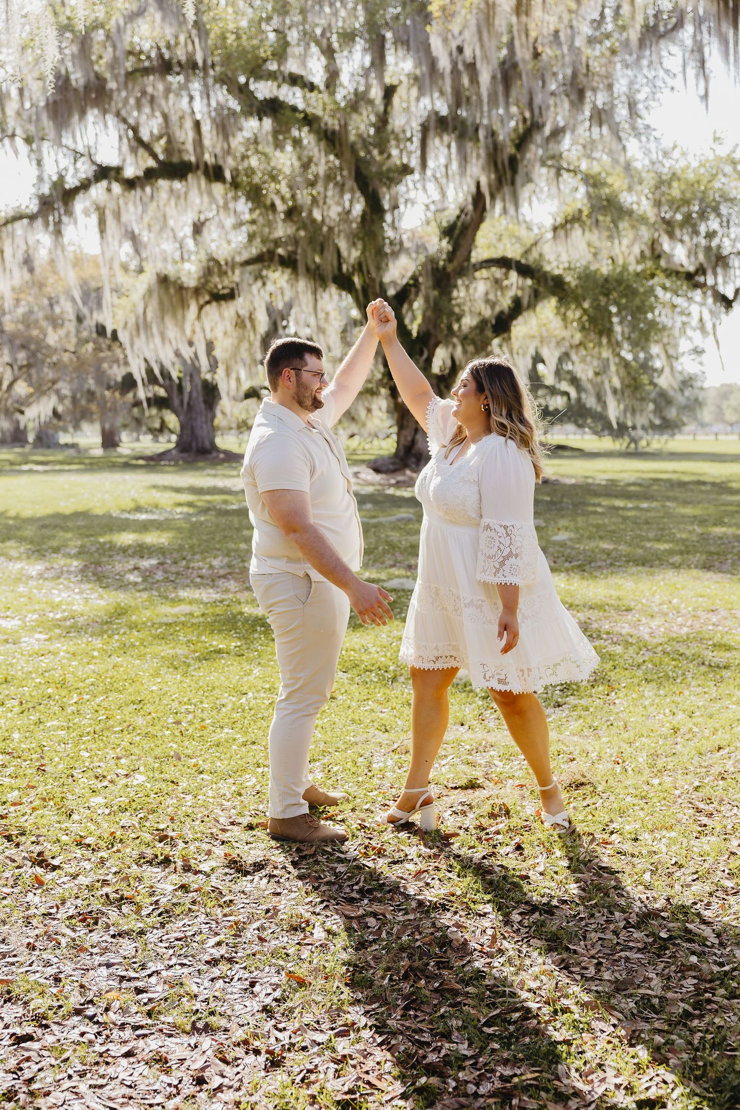 Couple in white outfits dancing under Spanish moss-draped oak tree in sunny park.