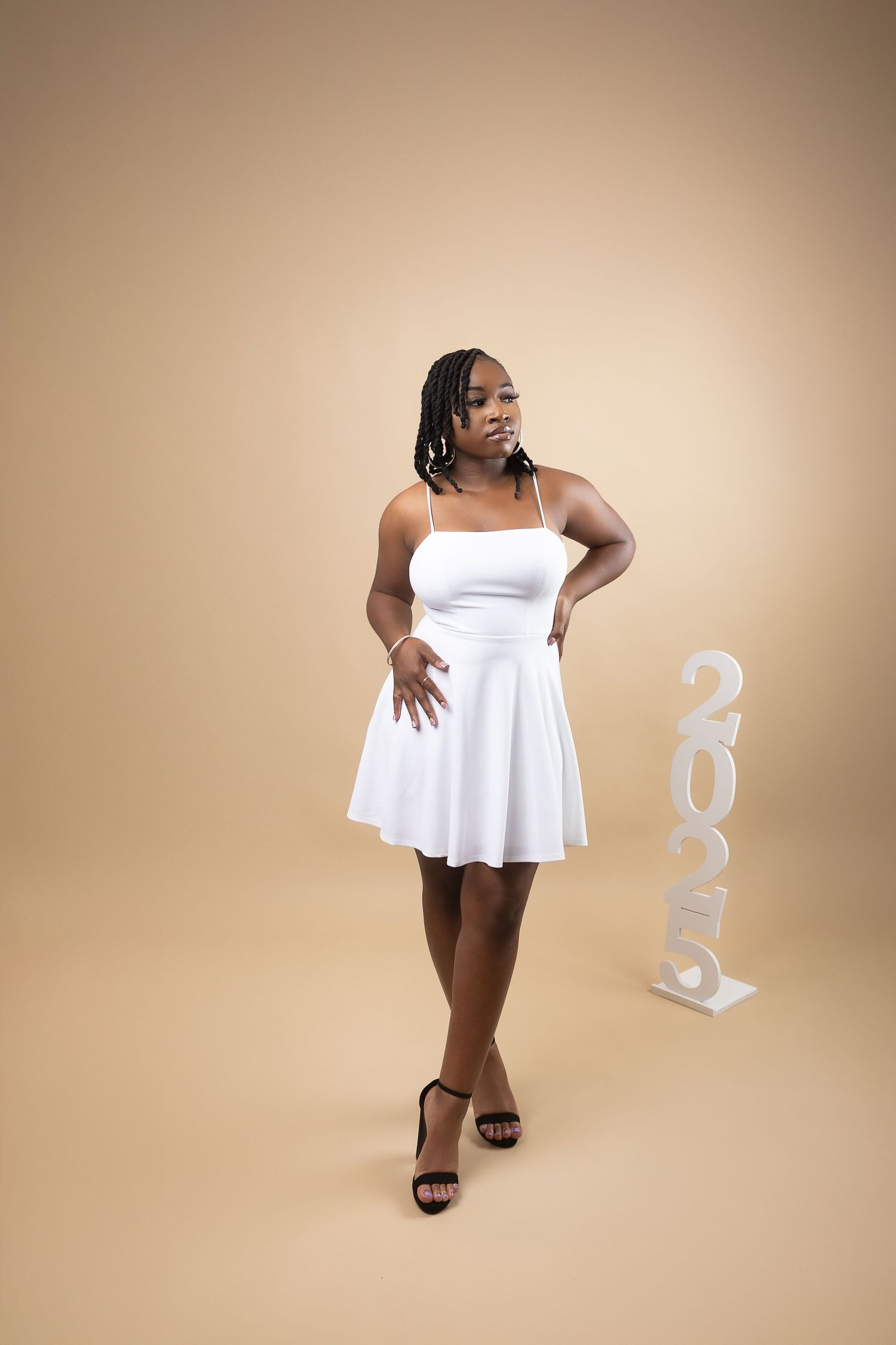 Young woman in white dress posing elegantly beside 2025 sign on warm beige background.