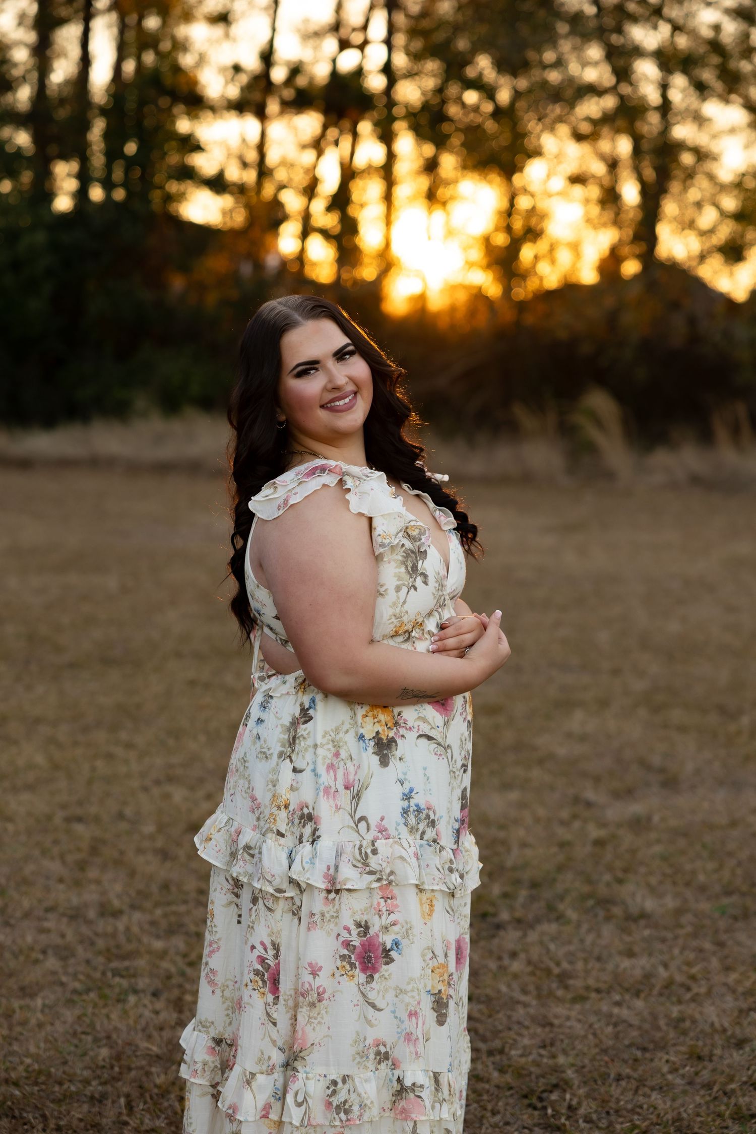 Smiling young woman in floral dress posing at golden sunset in outdoor field portrait.