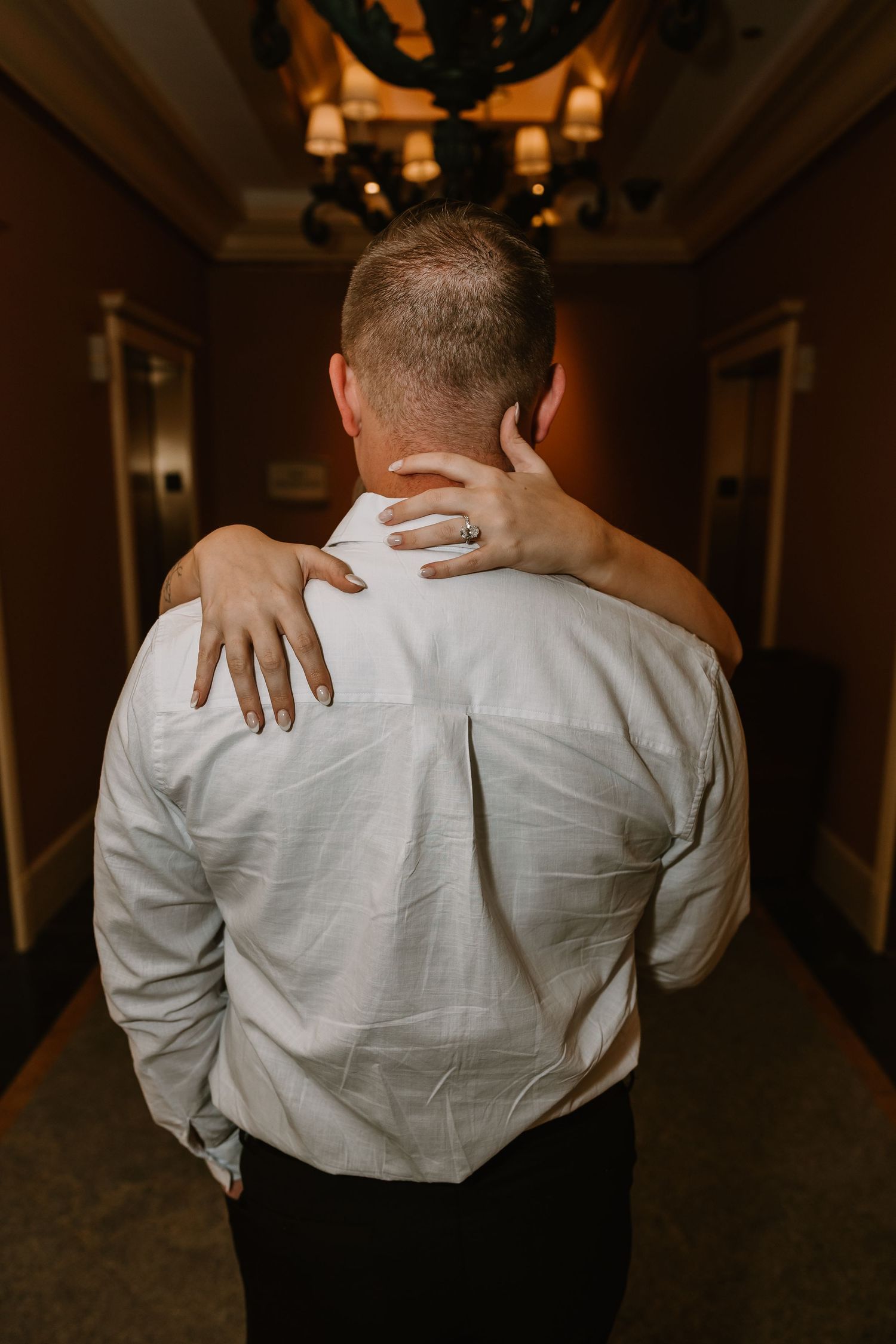Newly engaged woman with diamond ring rests hand on mans shoulder in elegant hotel corridor.