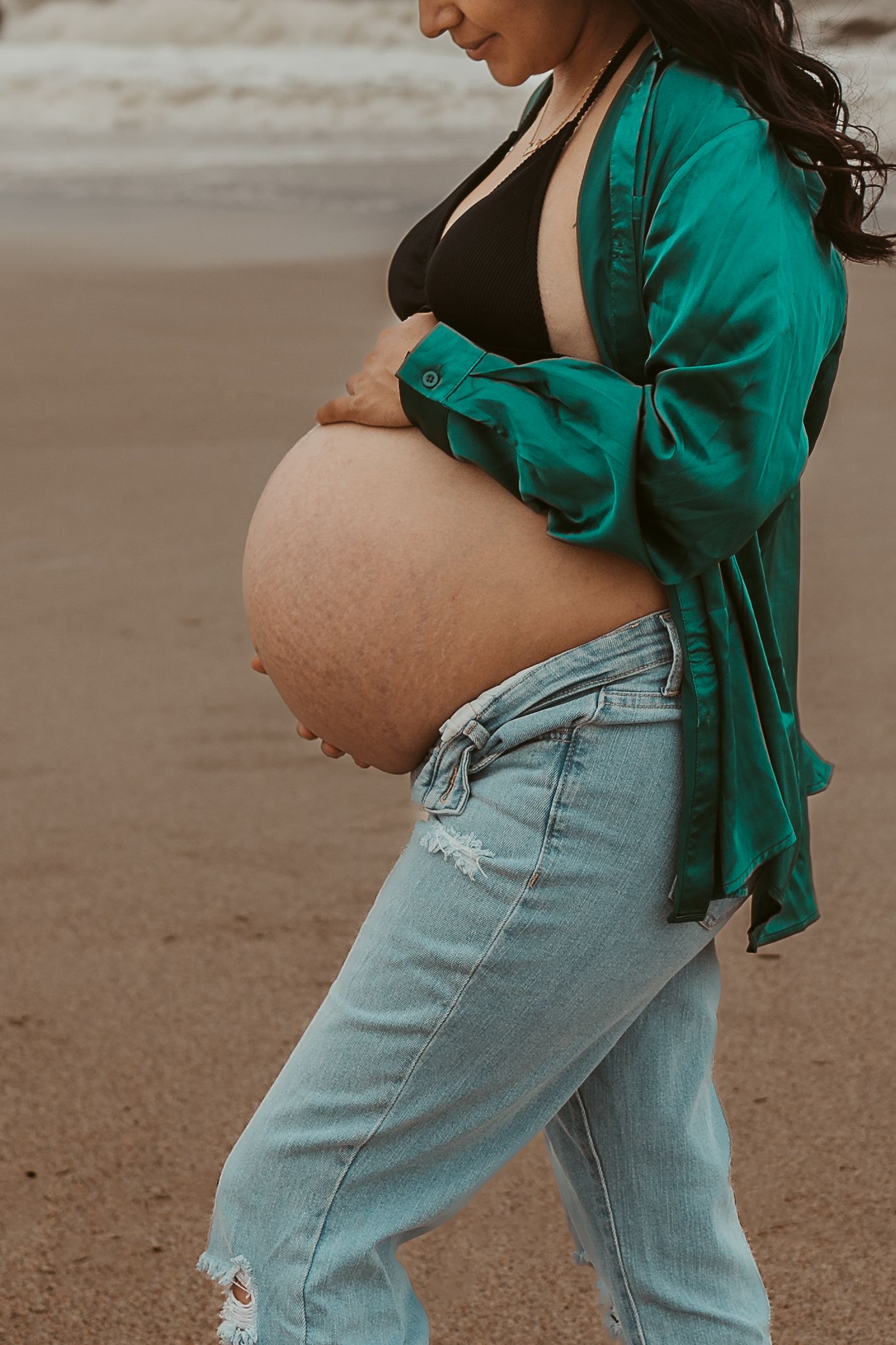 Close-up of pregnant womans bare baby bump wearing green shirt and ripped jeans at beach.