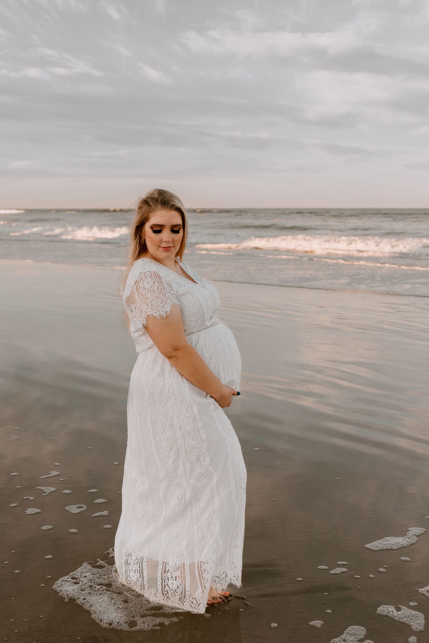 Smiling pregnant woman in white lace dress standing in ocean waves on beach at sunset.