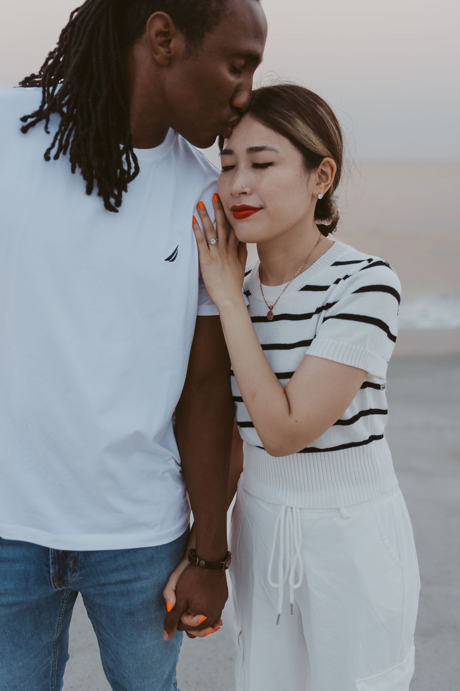 Interracial couple sharing a tender moment on the beach, man kissing woman's forehead while holding hands.
