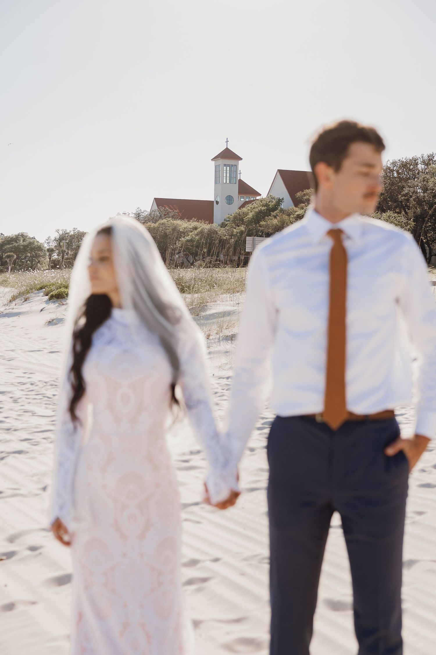 Bride and groom hold hands on beach with a charming chapel visible in background.