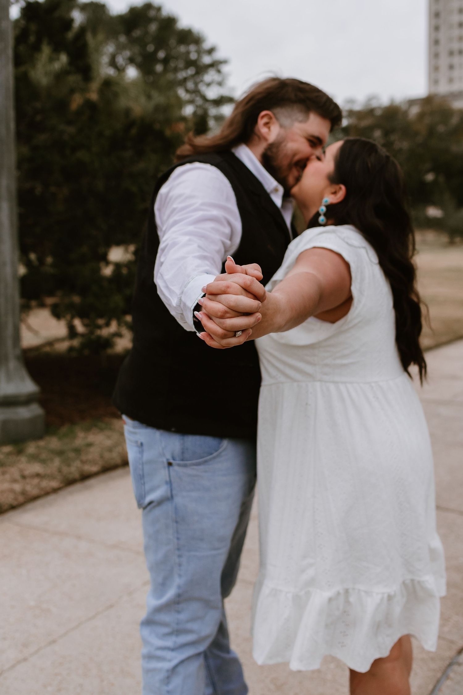 Couple sharing a passionate kiss while dancing outdoors, man in black vest and jeans, woman in white dress.