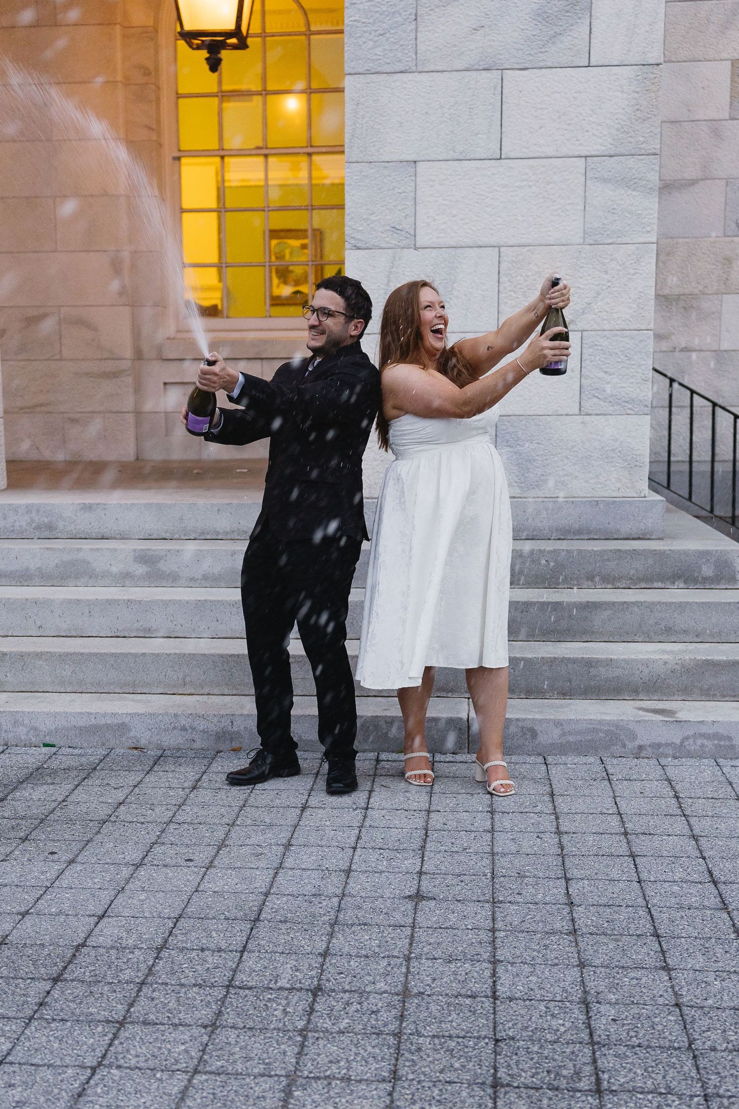 Joyful newlyweds popping champagne bottle on steps outside elegant building after wedding ceremony.