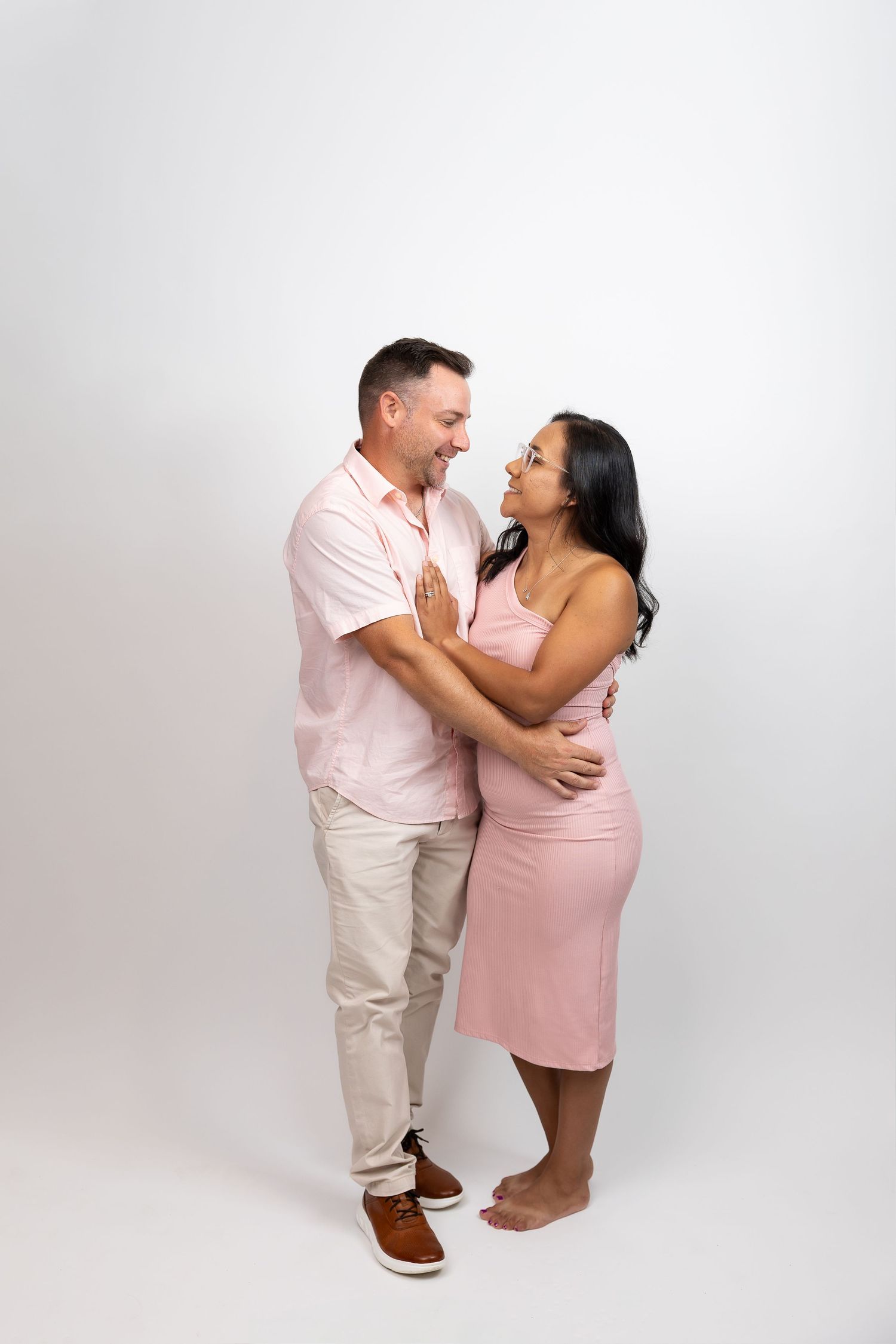 Smiling couple embracing during photo shoot, woman in pink dress on white background.