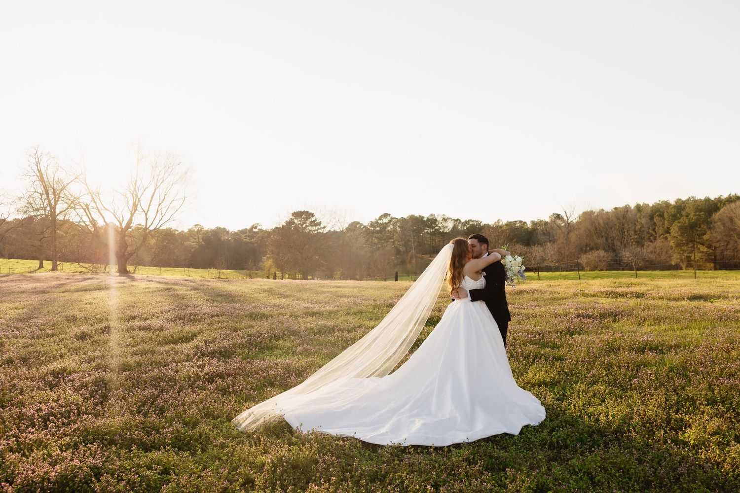 Bride and groom share a kiss in golden sunset light on a serene open field with flowing veil.