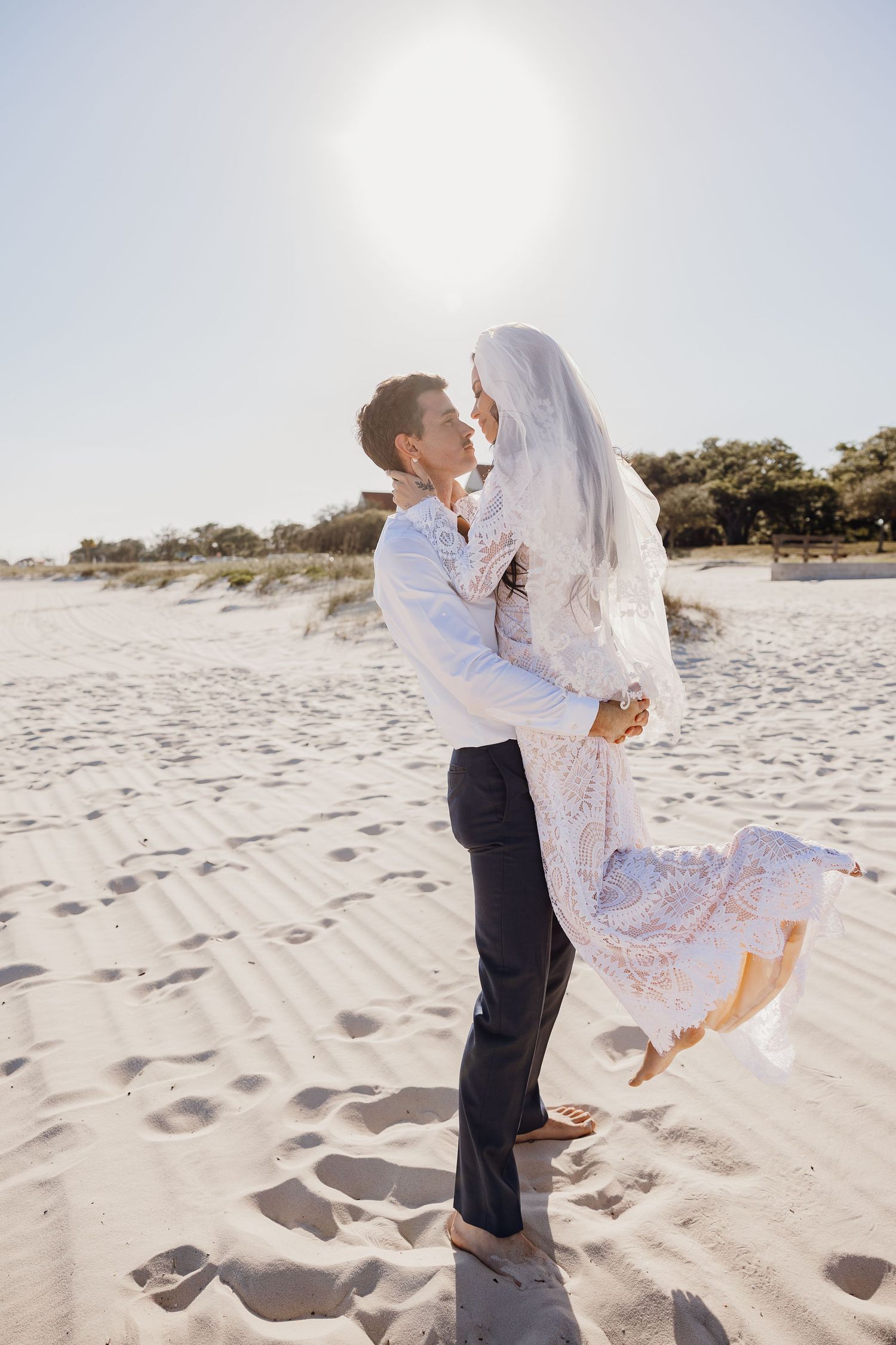 Groom lifting bride in white lace dress and veil on sandy beach in bright sunlight.