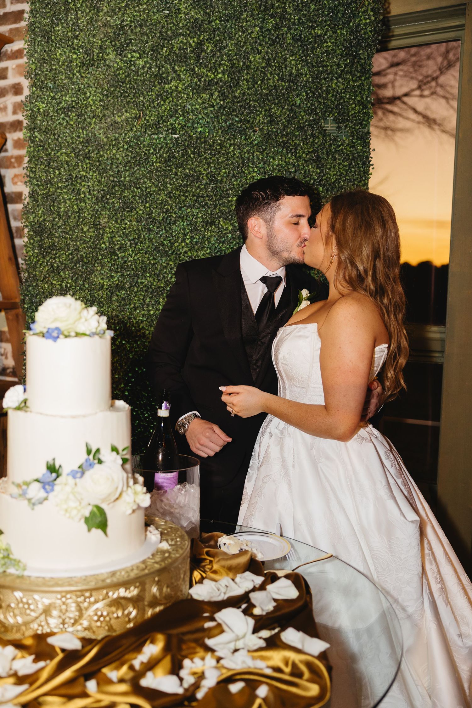 Bride and groom share a kiss while cutting white tiered wedding cake at golden hour reception.