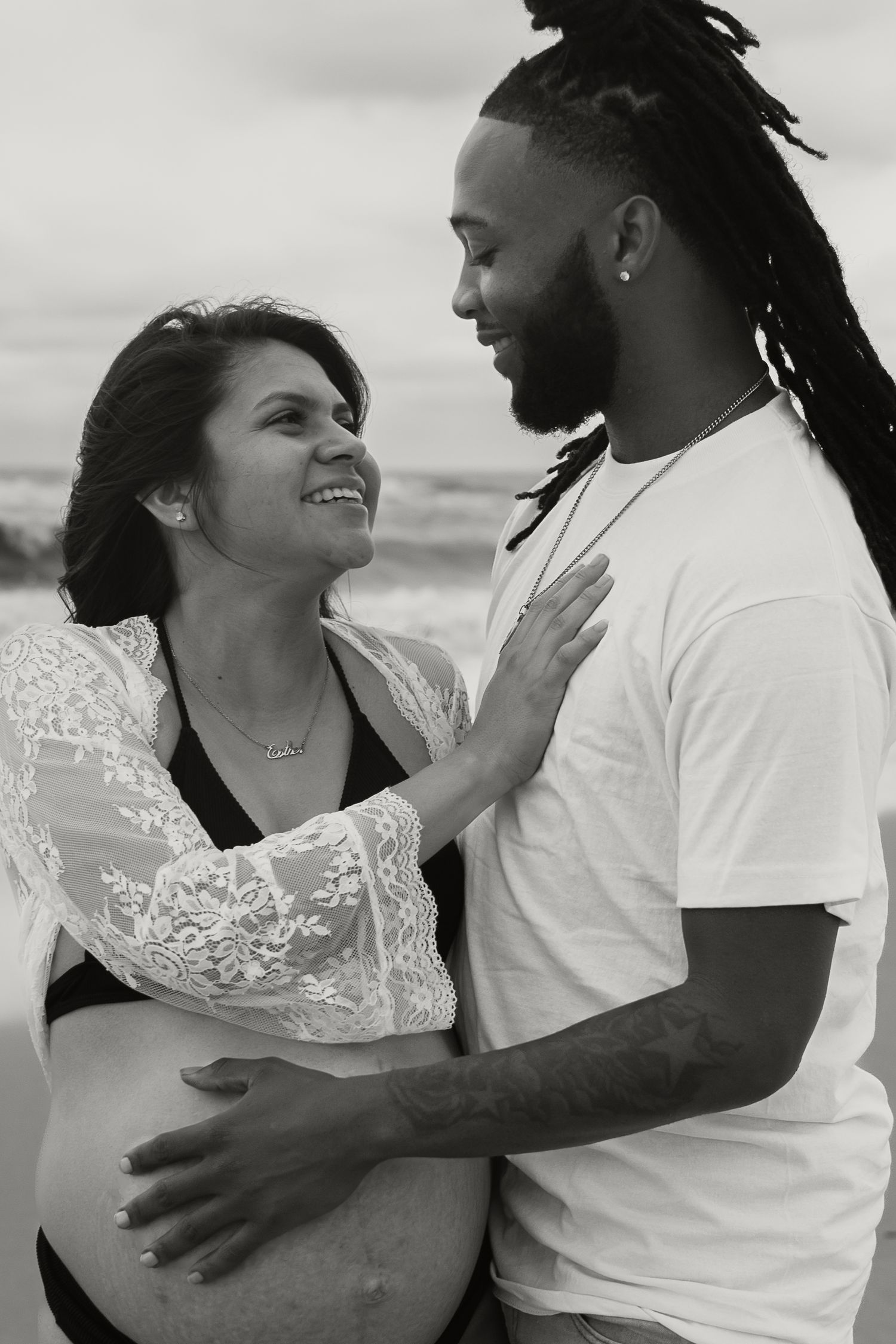 Black and white photo of interracial couple embracing on beach, man touching pregnant belly.