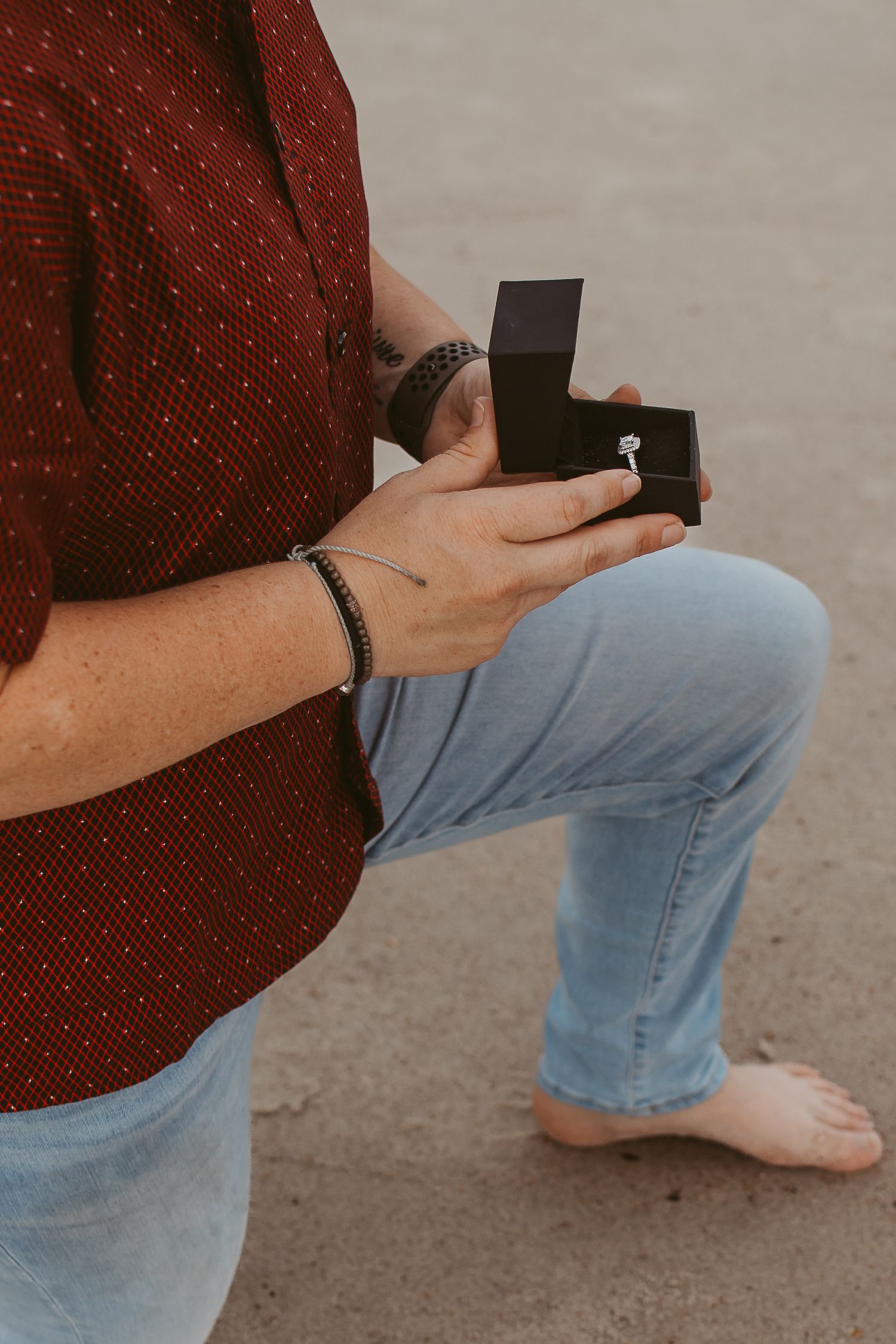 Close-up of person kneeling on beach holding open ring box with diamond engagement ring inside.