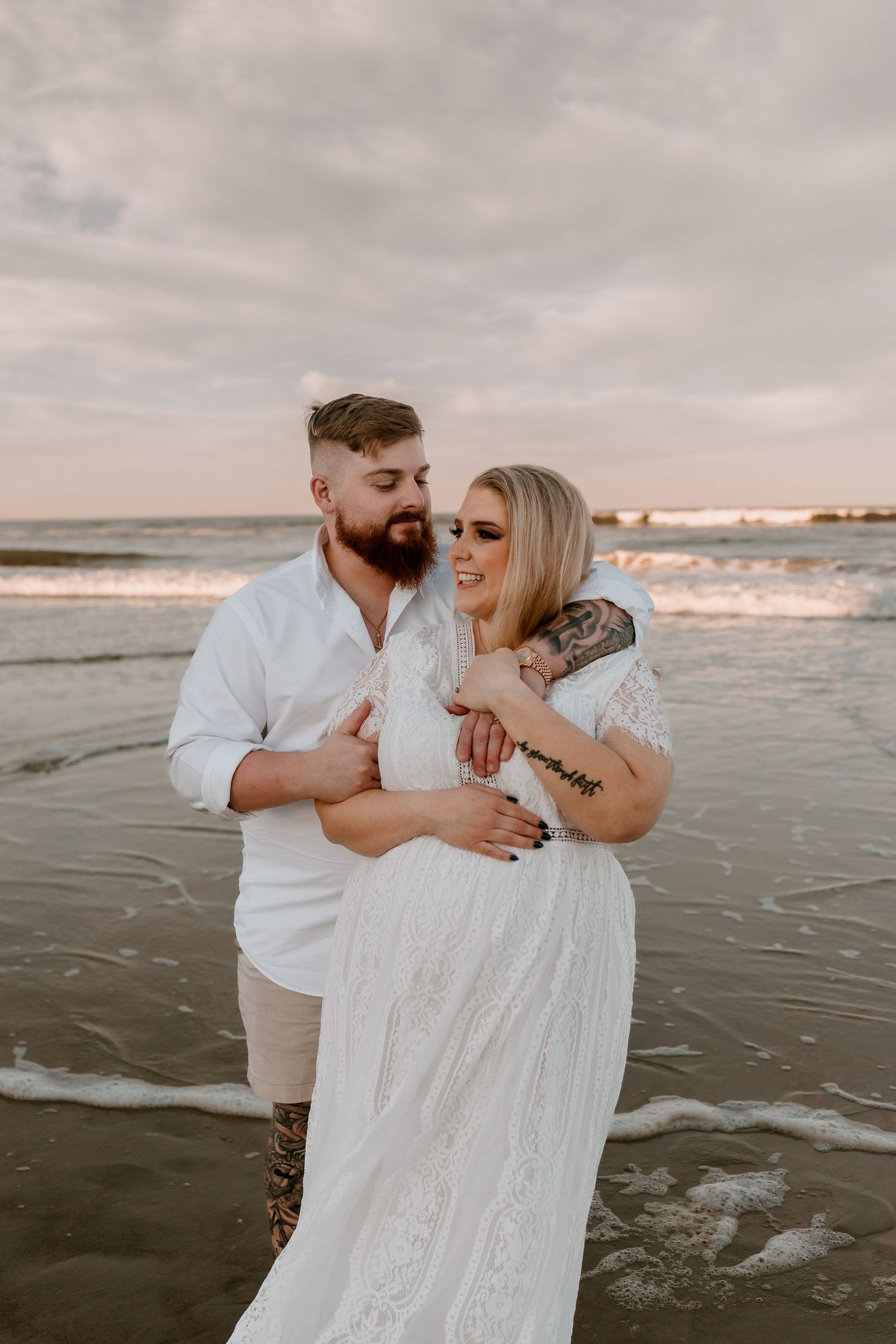 Tattooed couple embracing on beach at sunset, man in white shirt hugging pregnant woman in white lace dress.