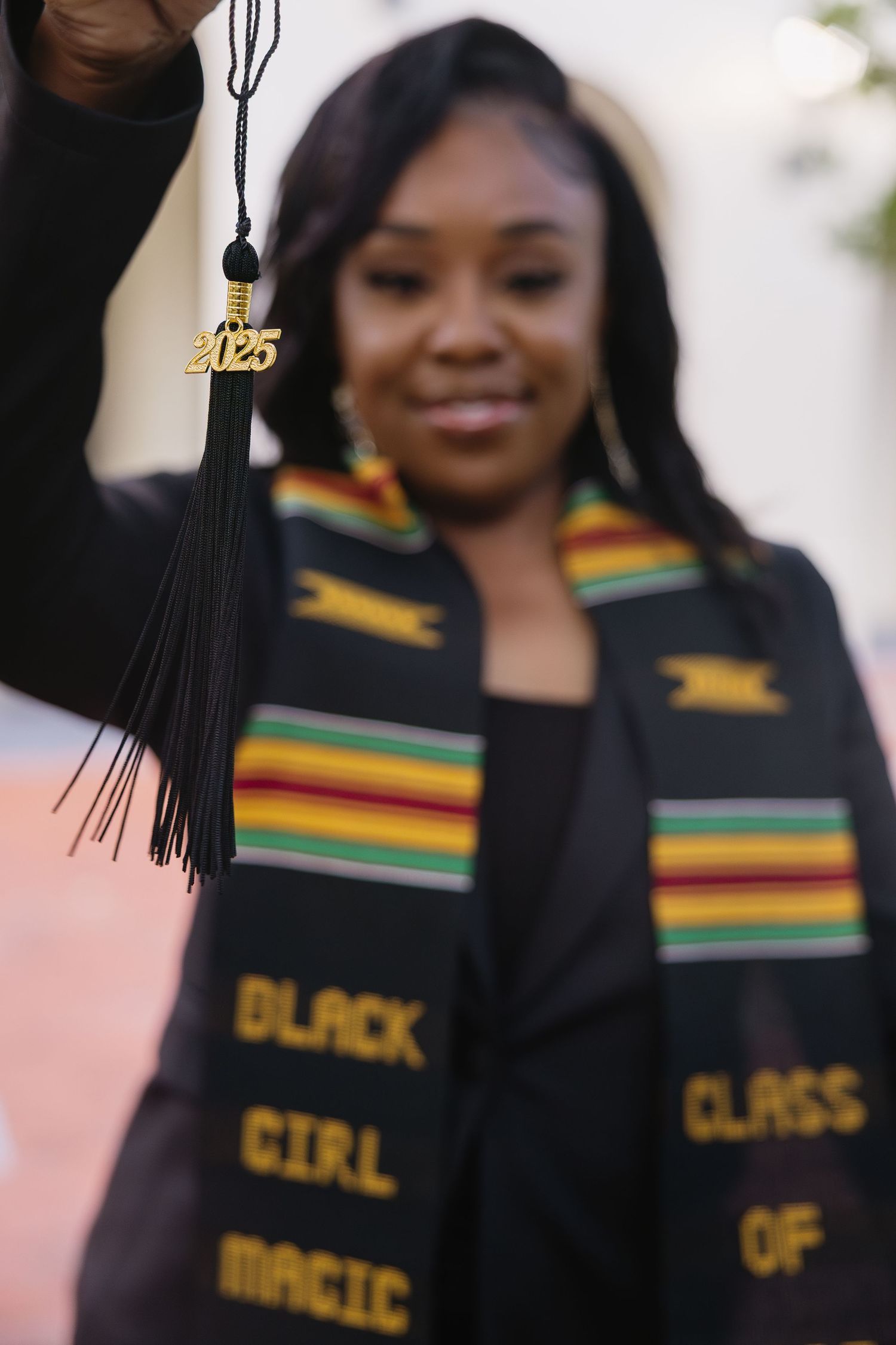A graduate holds up a 2025 tassel wearing a Black Girl Magic kente stole on campus.