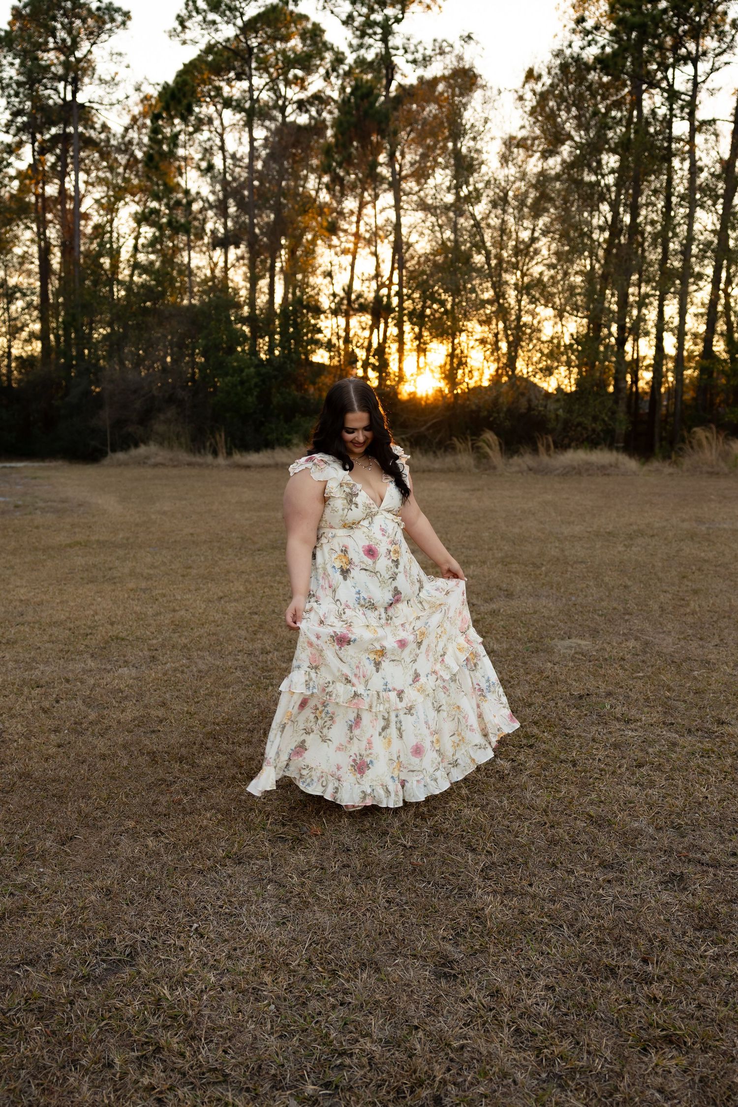 Brunette woman in floral dress looking down while spinning at sunset in field.