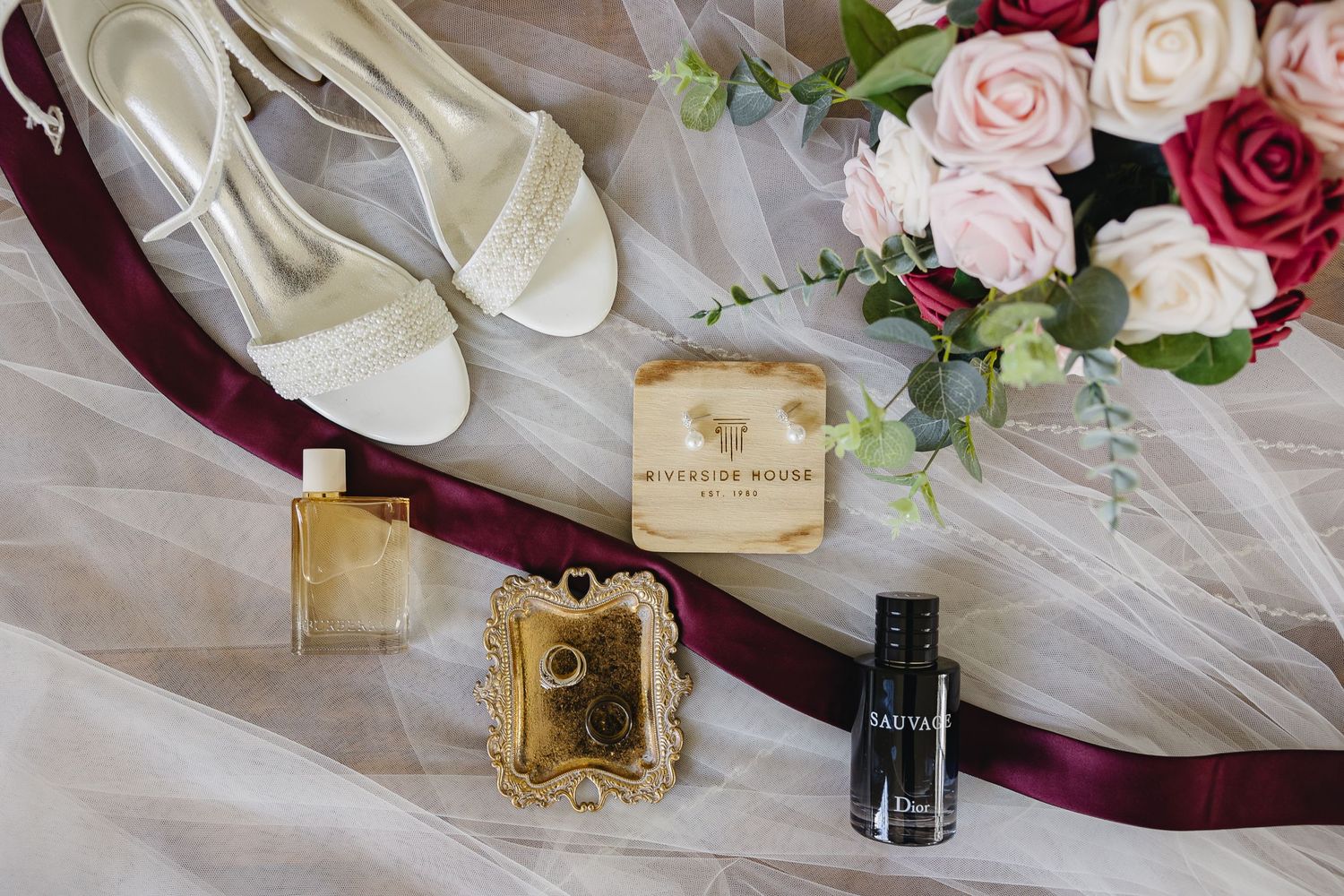 Bridal flat lay featuring white heels, perfume, Dior nail polish, and floral bouquet on veil.