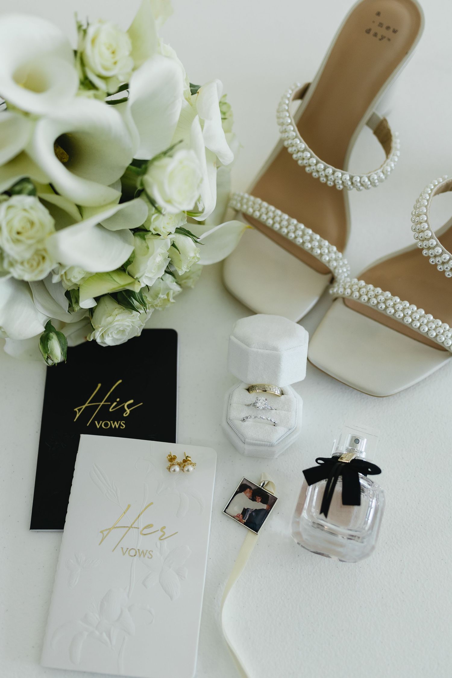 Bridal flat lay with pearl heels, white bouquet, vow books, ring box, and perfume.