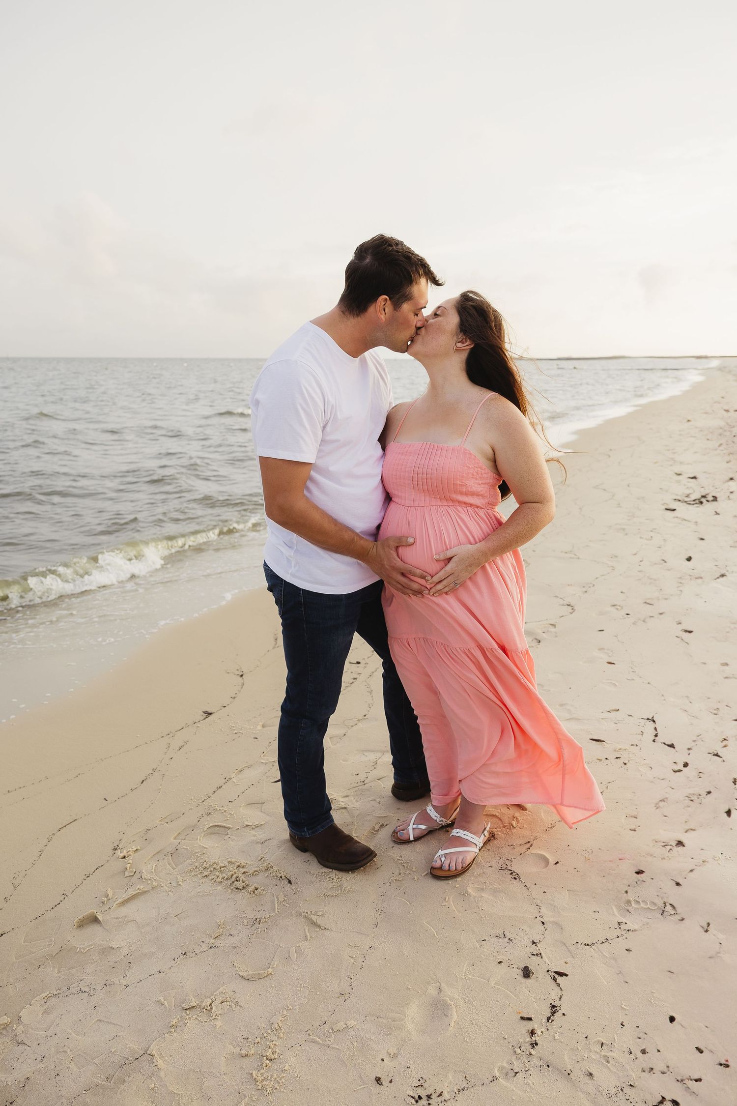 Pregnant couple kissing on white sand beach at sunset, woman in pink dress holding baby bump.