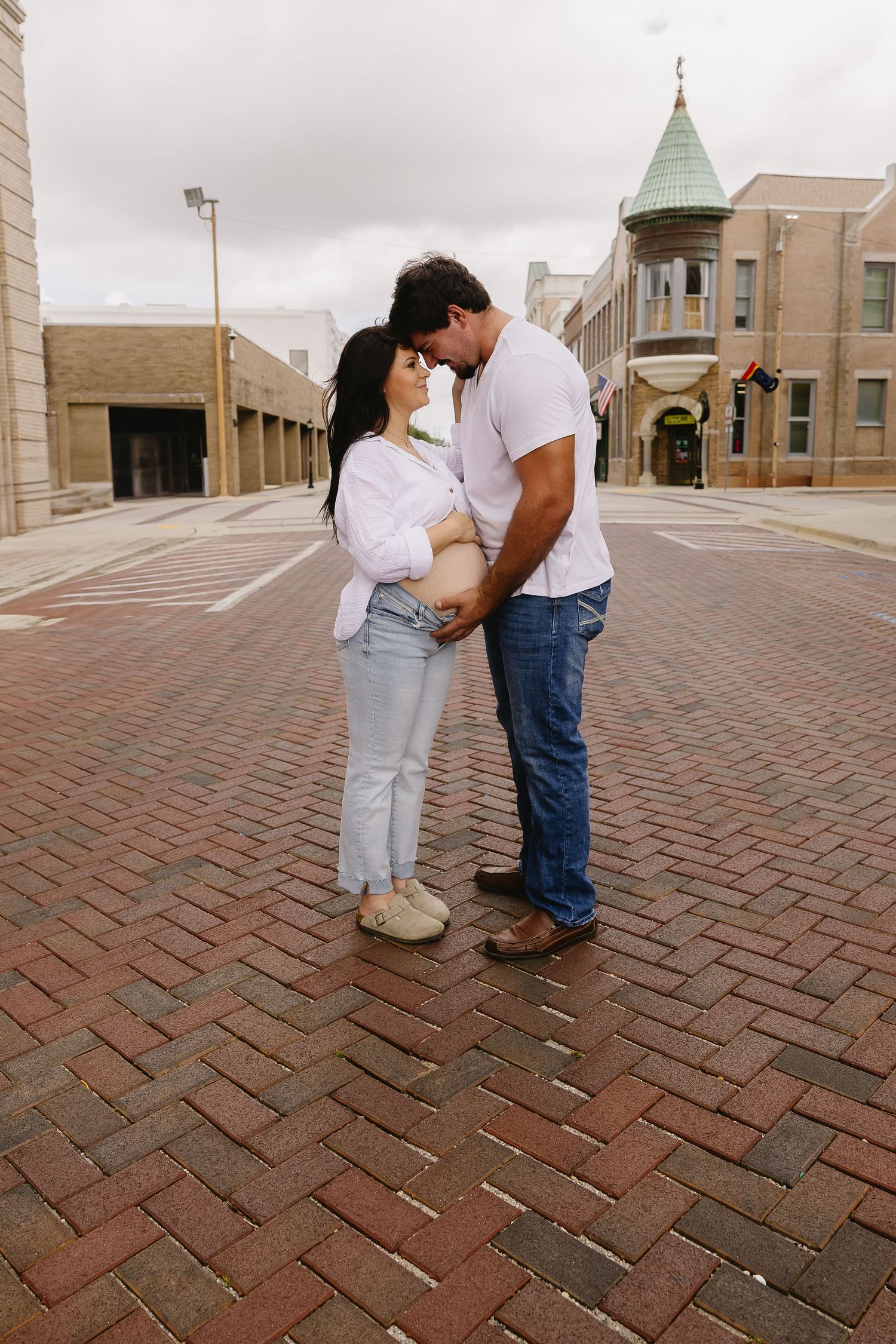 Expecting couple touching foreheads and holding baby bump on a brick street in an urban setting.