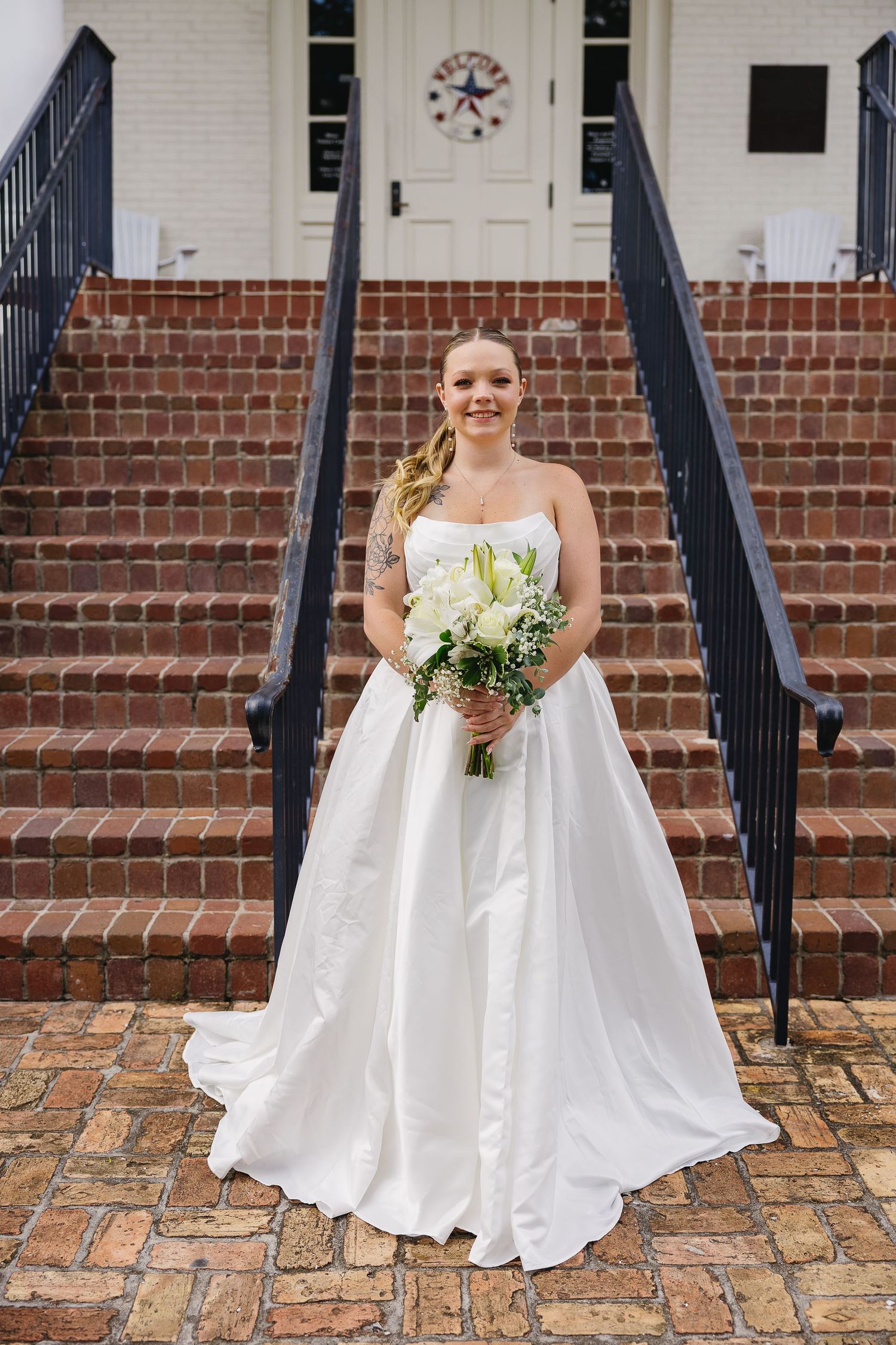 Bride in strapless white ballgown holding bouquet on brick steps of a white chapel.