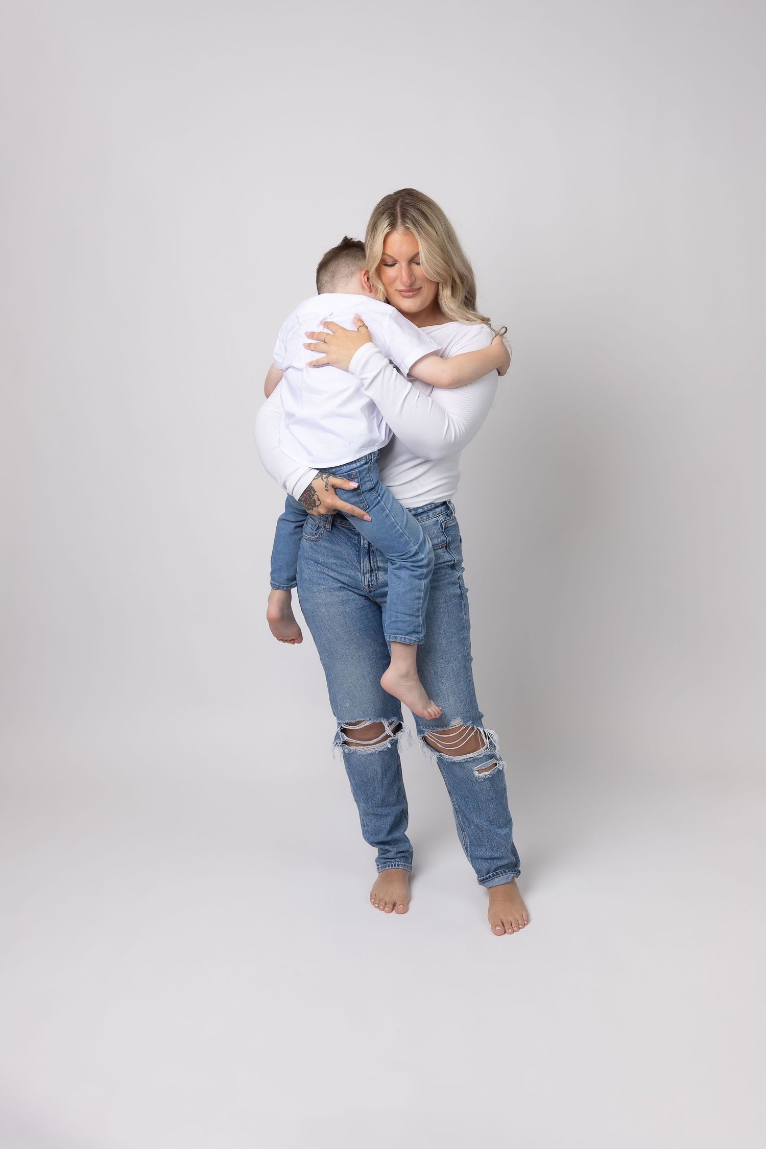 A blonde mother in ripped jeans hugs her young child in a clean white studio photo.