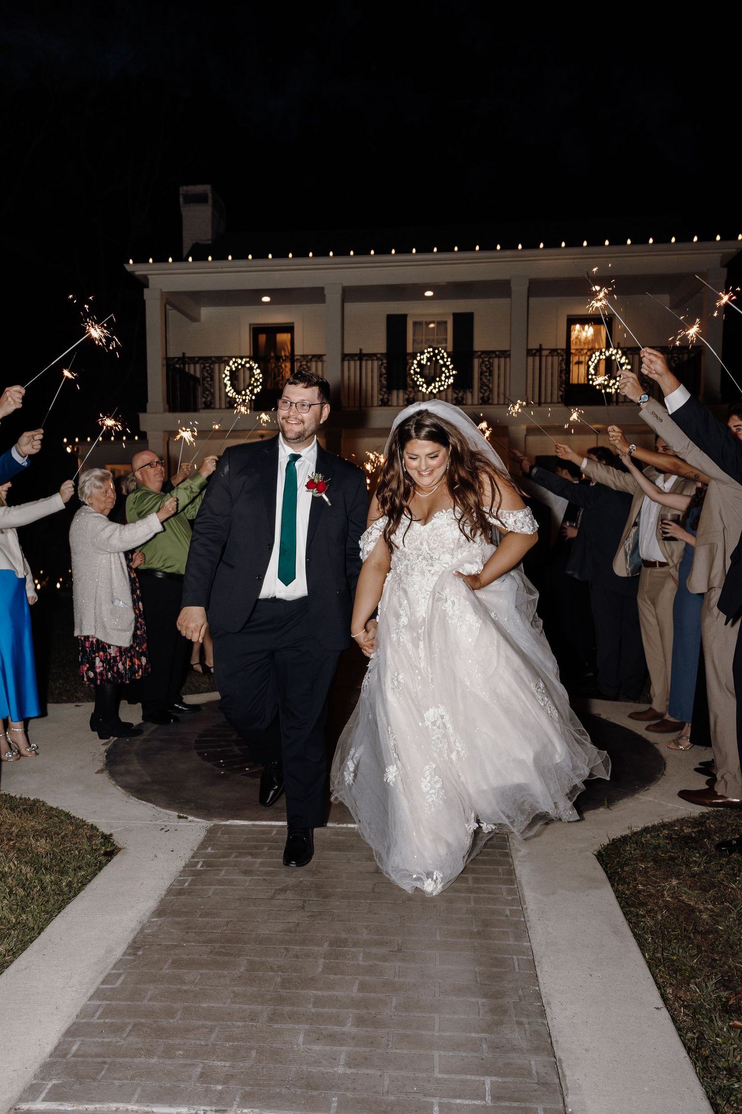 Newlyweds exit through sparkler tunnel held by guests outside a decorated white venue at night.