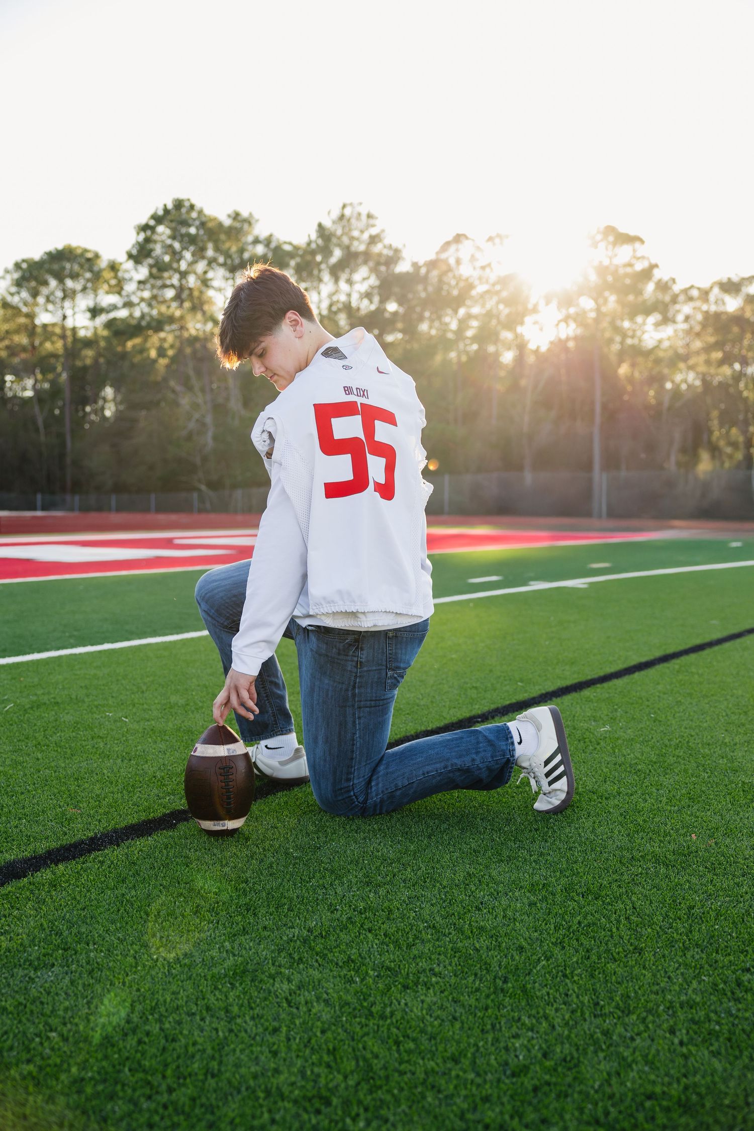 Teen in white number 55 hoodie kneeling with football on green turf at sunset.