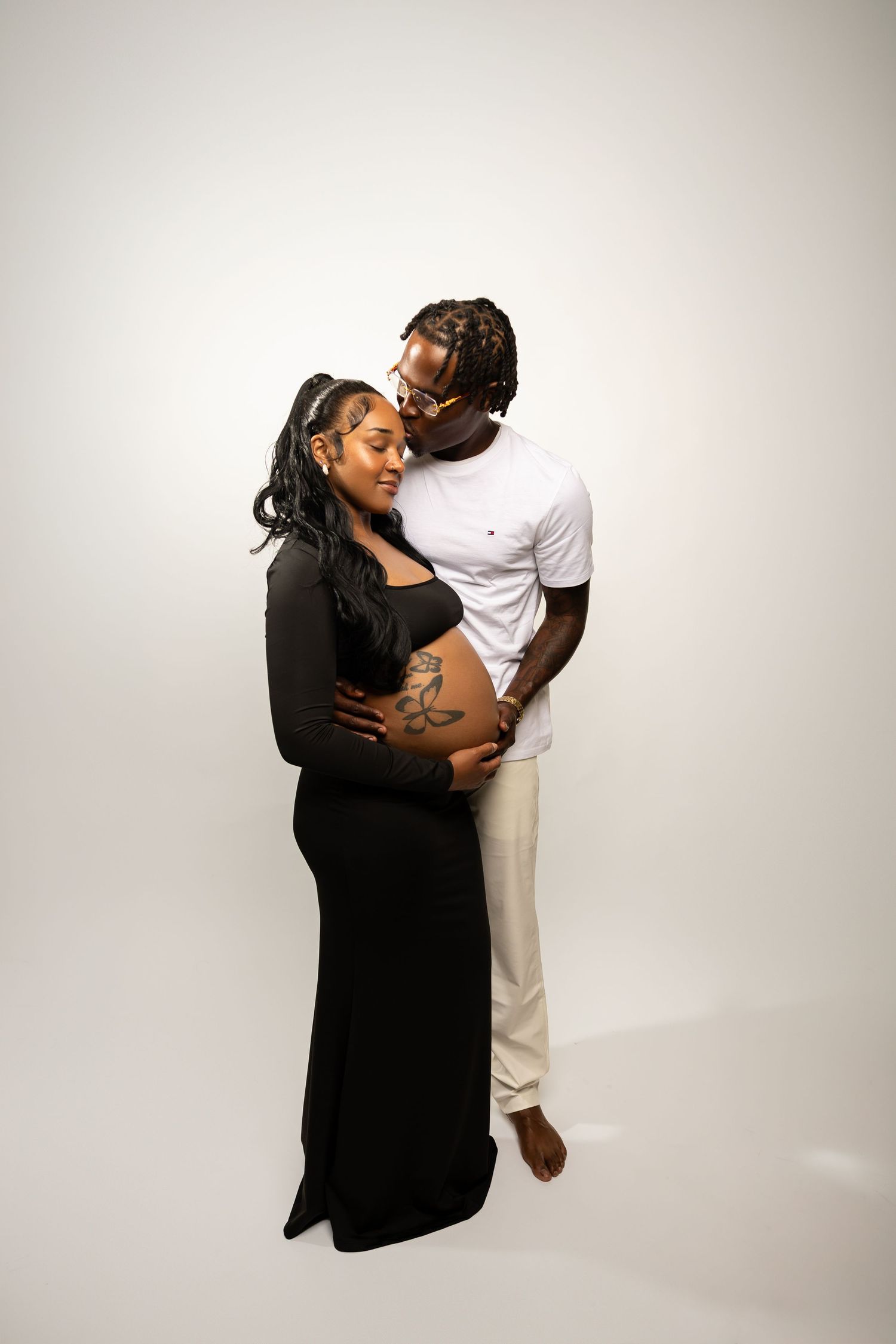 Couple embraces tenderly as man kisses pregnant woman's tattooed baby bump on white backdrop.