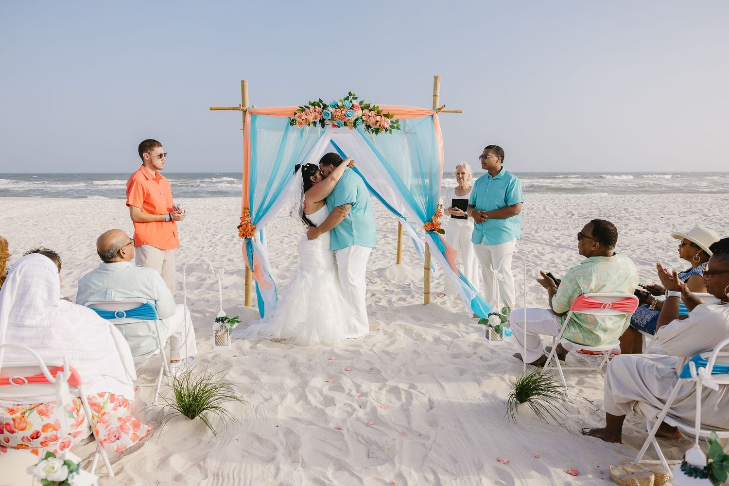 Couple shares first kiss under turquoise-draped bamboo arch at intimate beach wedding ceremony.