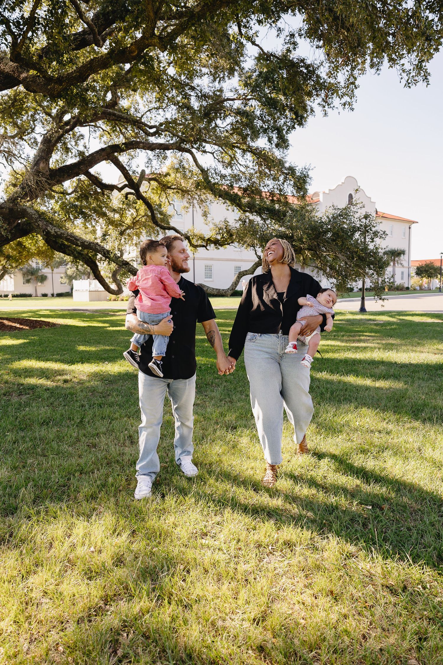 Young family of four walking hand in hand under large oak tree at golden hour outdoors.